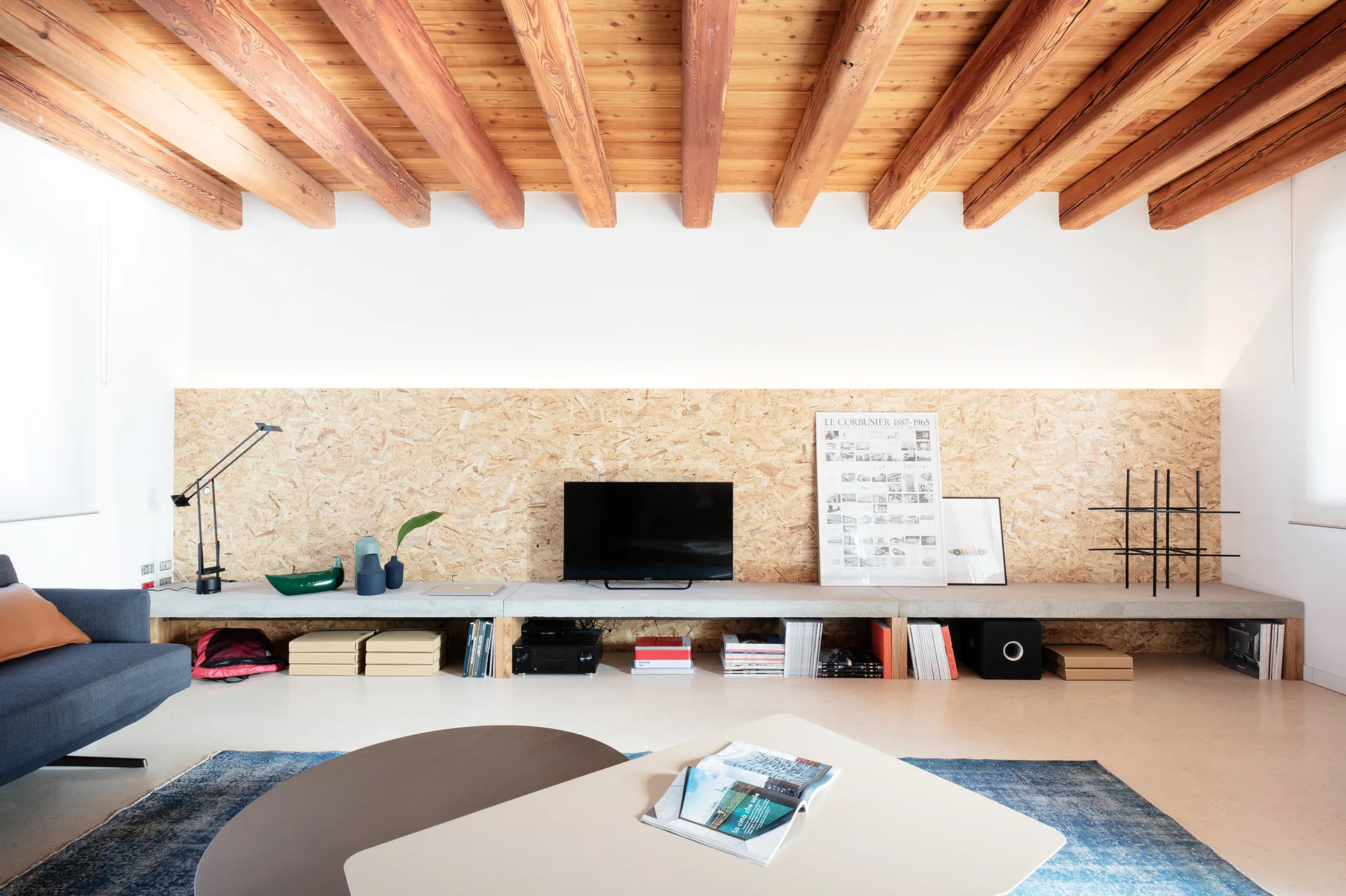 A modern living room features a low-profile concrete console below an OSB wood panel wall, with exposed wooden ceiling beams overhead.