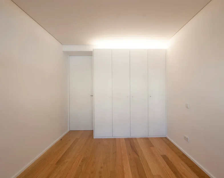 An empty minimalist bedroom features light wood floors, a flush white door, and a built-in floor-to-ceiling wardrobe highlighted by overhead cove lighting.