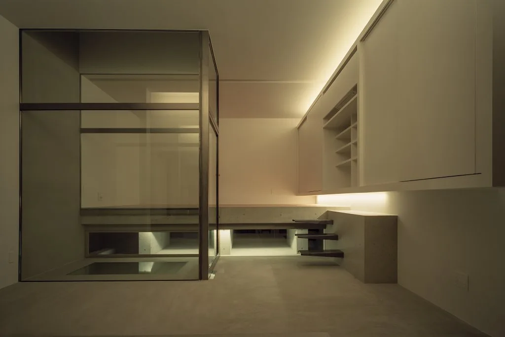 A minimalist room features a large glass-walled enclosure, a raised concrete platform with small floating stairs, and warm under-cabinet lighting.
