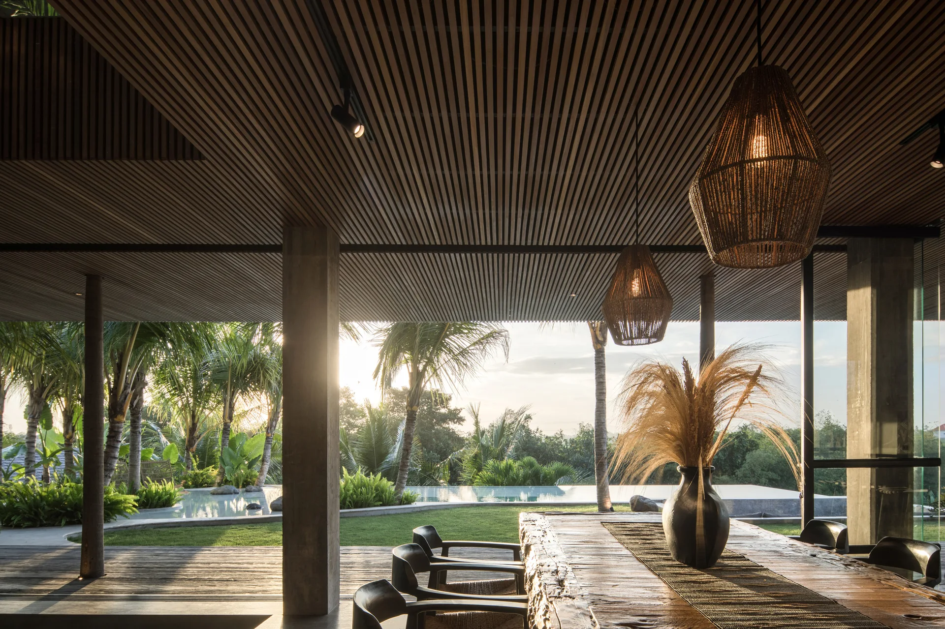 A covered outdoor dining patio features a rustic wood table beneath a slatted ceiling, overlooking a backyard swimming pool and palm trees.