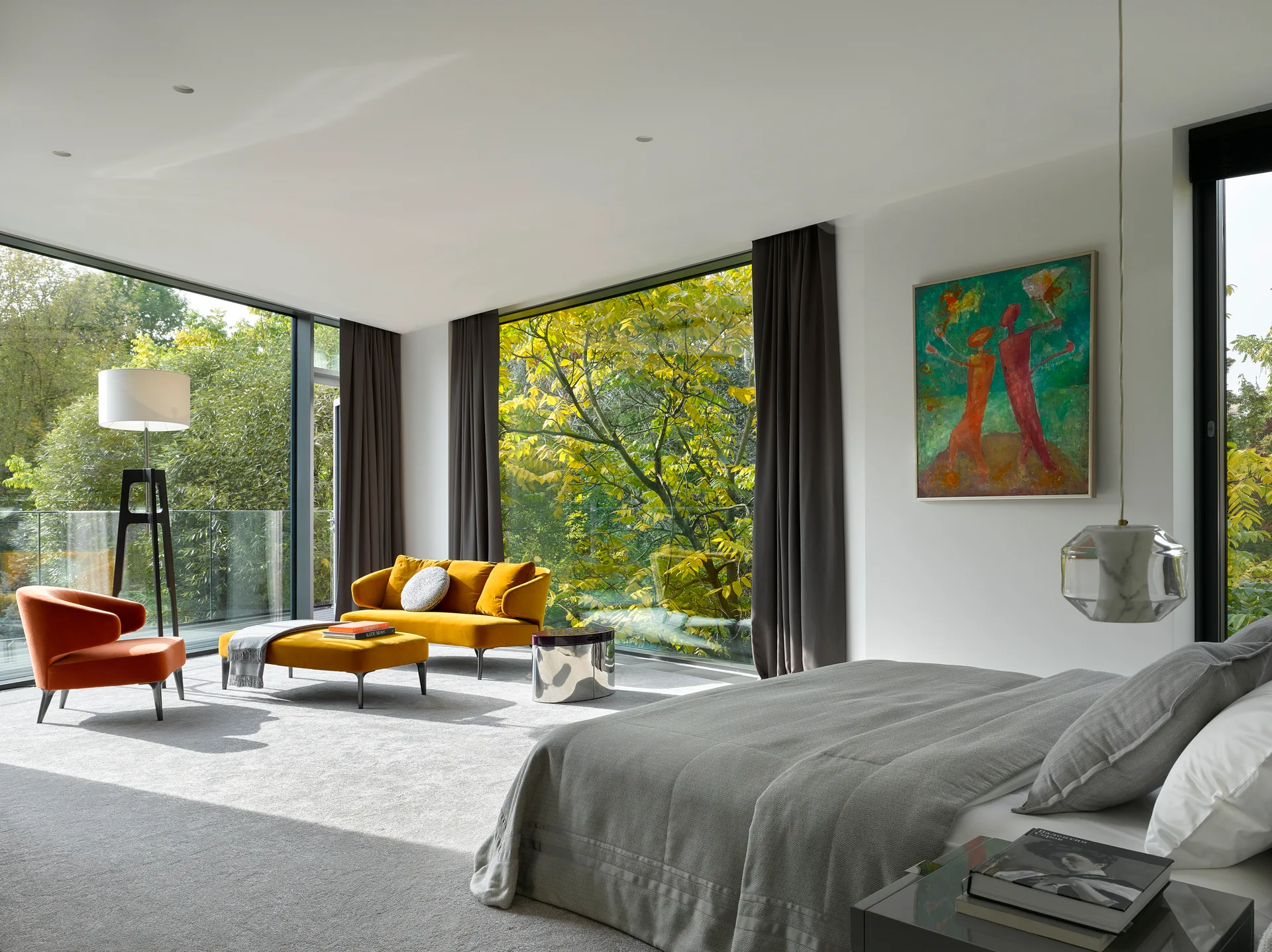 A modern bedroom featuring a grey bed, a yellow velvet seating area, and large floor-to-ceiling windows overlooking lush green trees.