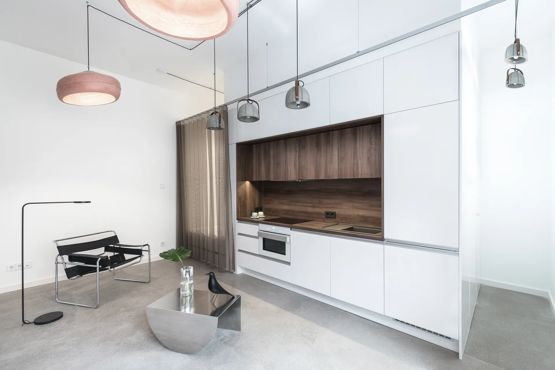 A minimalist open-plan kitchen featuring two-tone white and dark wood built-in cabinetry, a black leather armchair, and a polished metal side table.