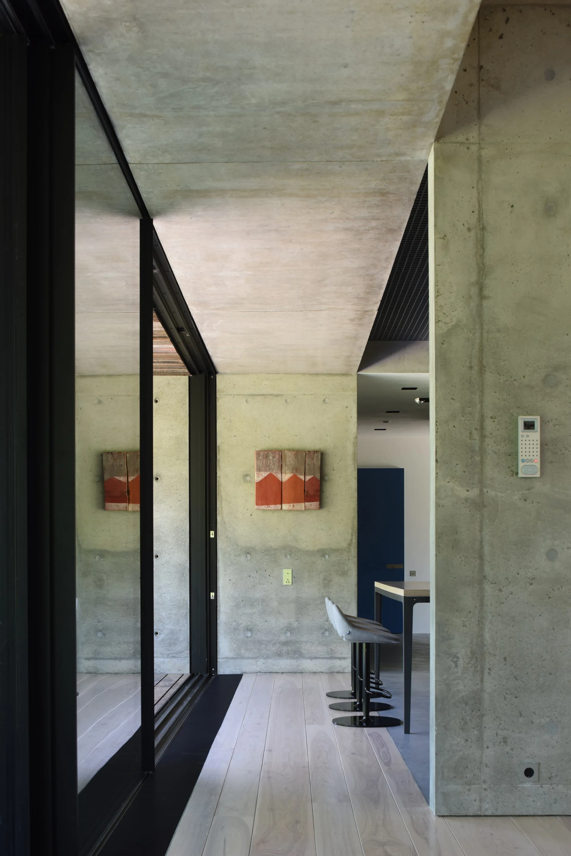 A modern residential hallway featuring exposed concrete walls, light wood plank flooring, and a large black-framed sliding glass door.