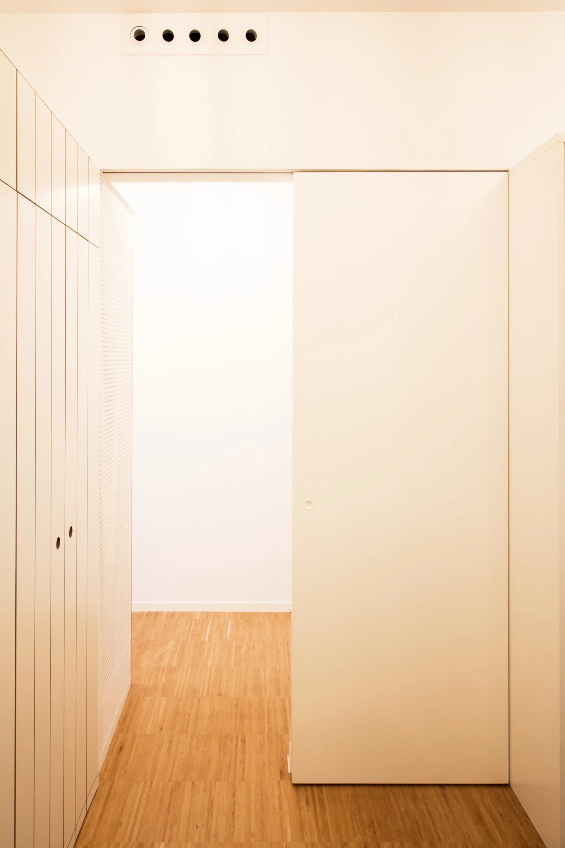 A minimalist hallway features light wood flooring, white floor-to-ceiling built-in cabinets with vertical grooving, and a large sliding door.