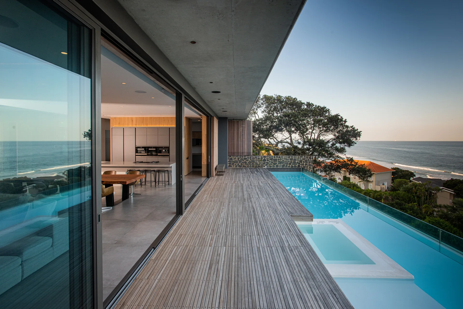 A modern concrete terrace features a wide wood deck, a bright blue infinity pool, and open sliding glass doors leading into an indoor kitchen.