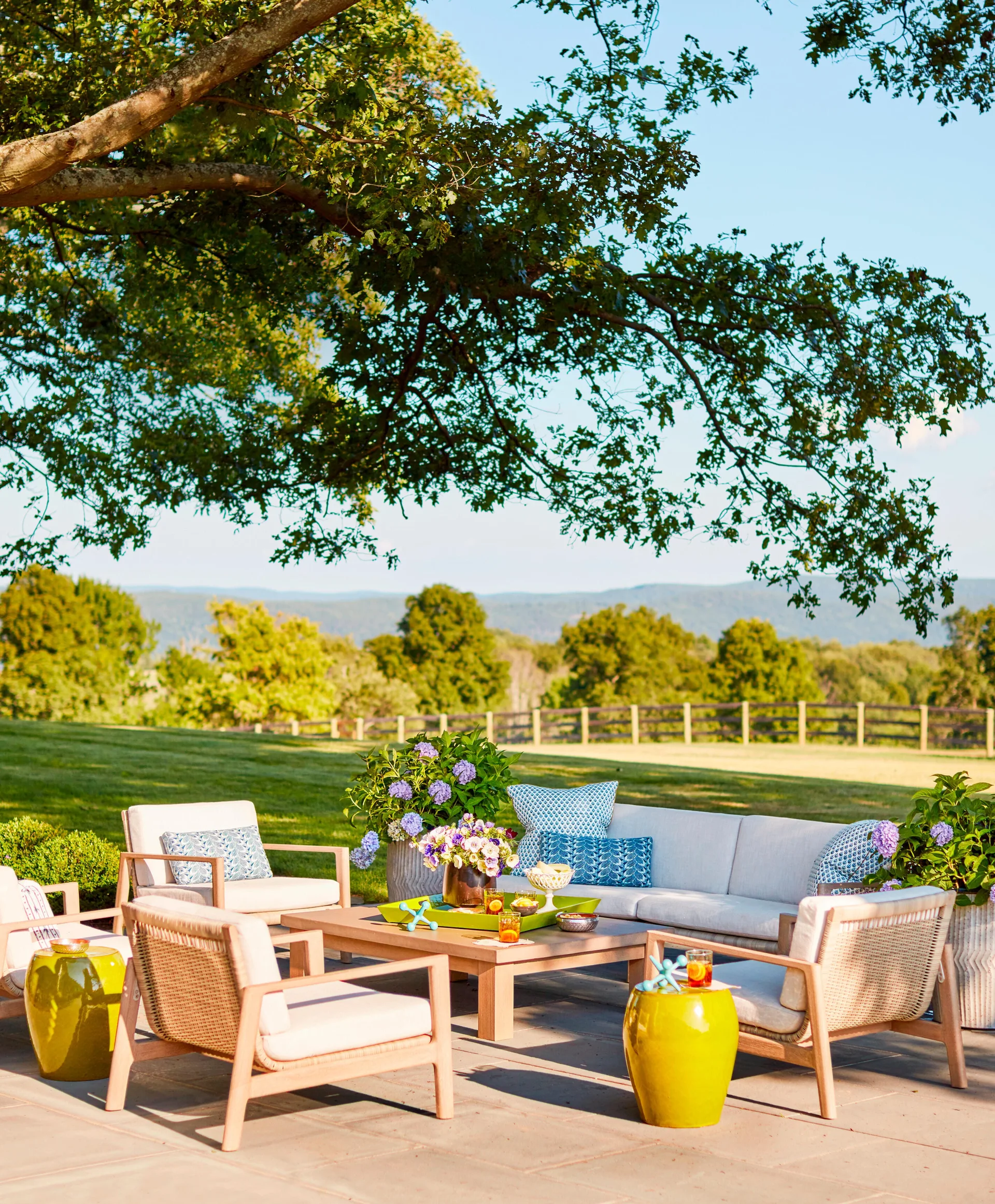 A stone patio under a shaded tree features a cream-cushioned wooden sofa, armchairs, blue patterned pillows, and bright green ceramic garden stools.