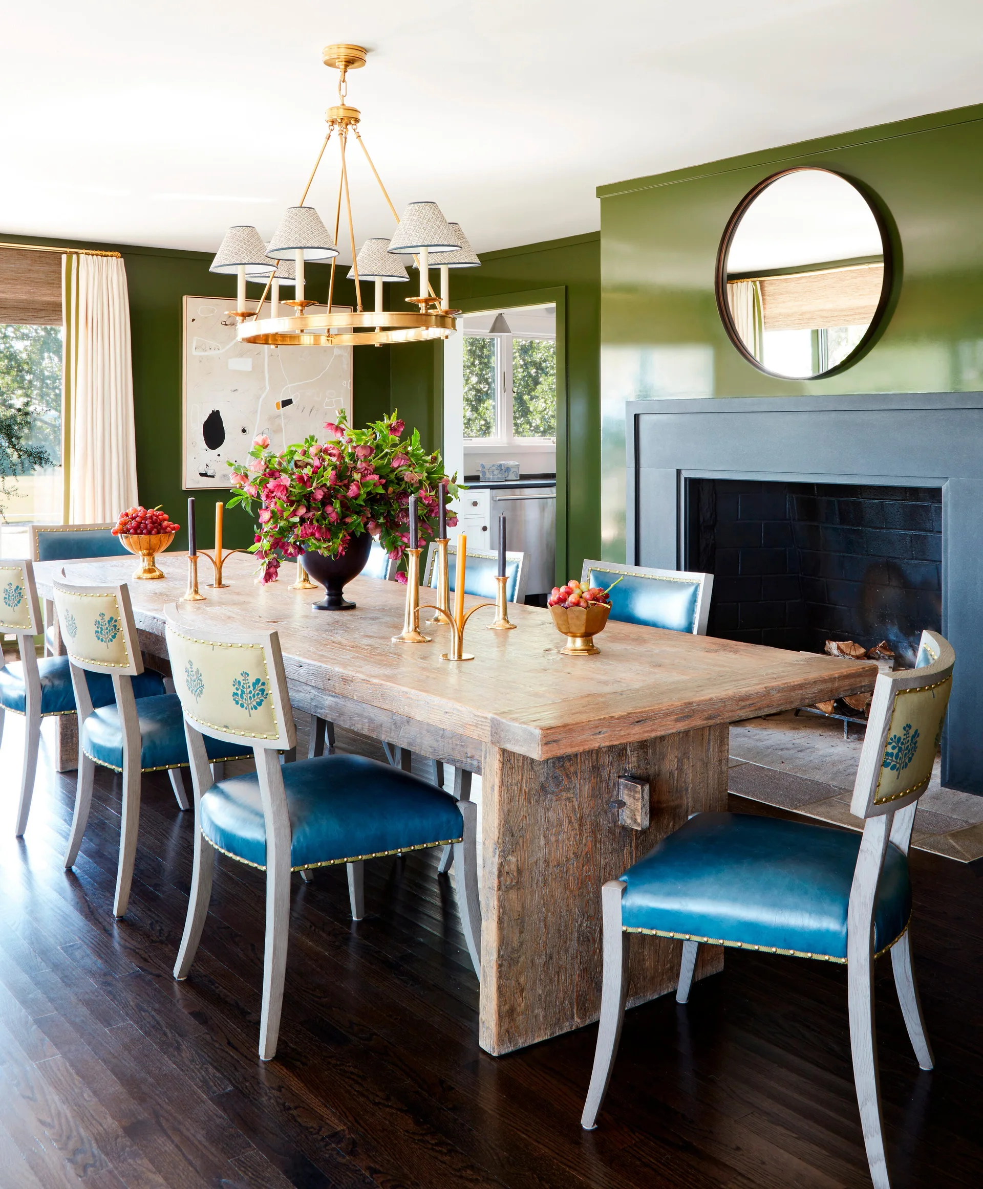 A rustic wooden dining table is surrounded by blue leather chairs in a room with glossy olive green walls and a dark fireplace.