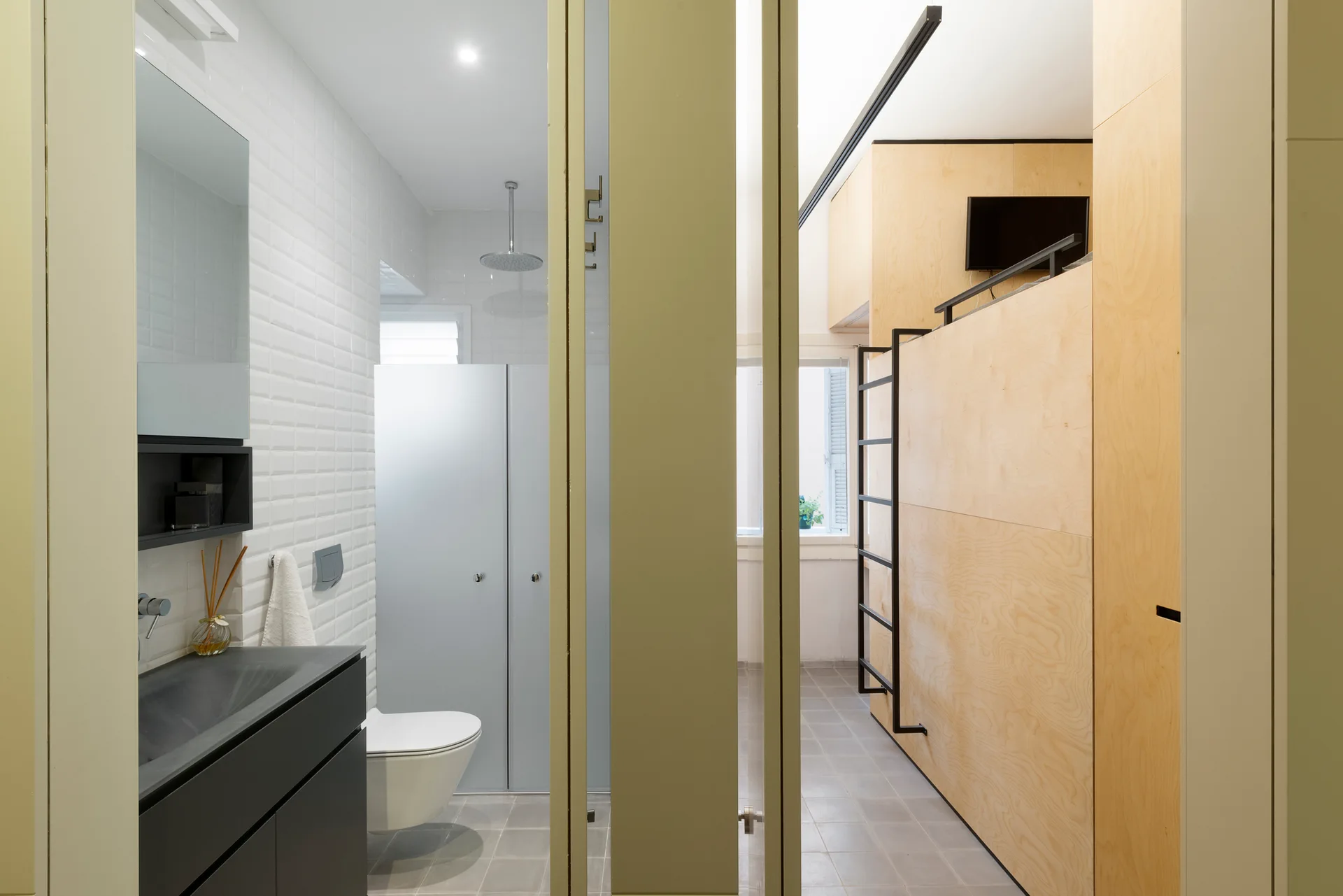 A modern bathroom with white subway tile walls and a black vanity sits next to a light plywood partition wall with an integrated black ladder.