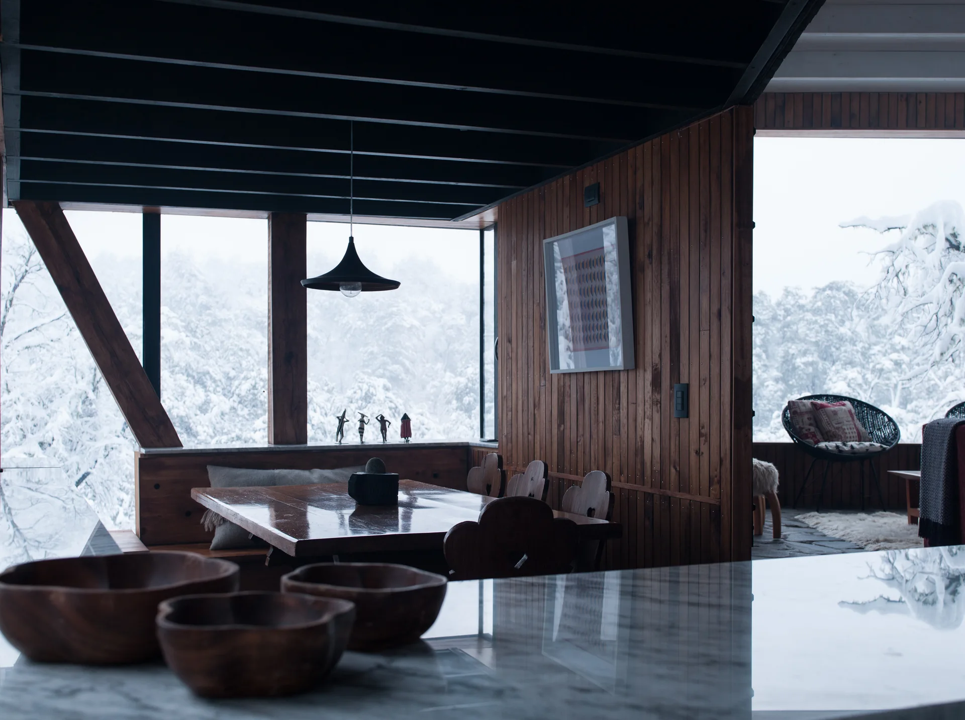 A rustic wood-paneled dining room features a wooden table and chairs under dark ceiling beams with a large window looking out at a snowy forest.