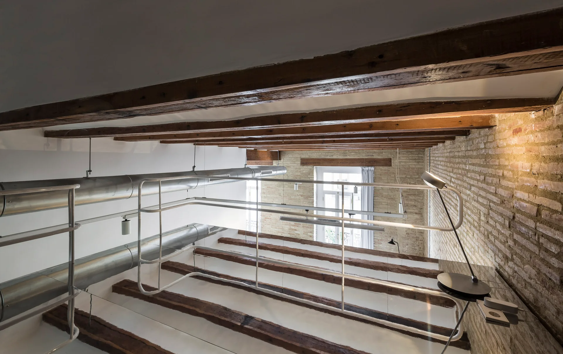 An industrial loft mezzanine features exposed dark wood ceiling beams, a raw brick wall, white metal railings, and silver ventilation ducts.
