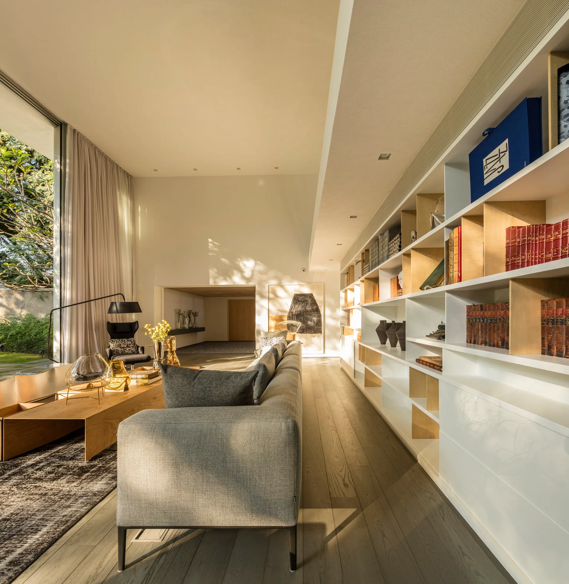 A modern living room features a long grey sofa, a low wooden coffee table, and a wall-length white bookshelf with light wood compartments.