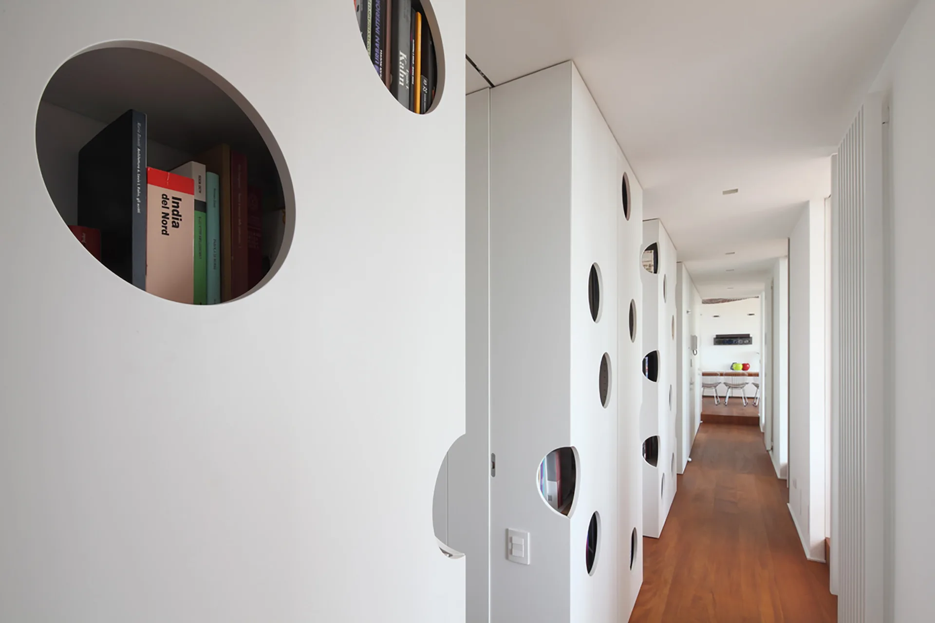 Minimalist white corridor with custom floor-to-ceiling storage units containing circular book niches alongside polished wooden floors.