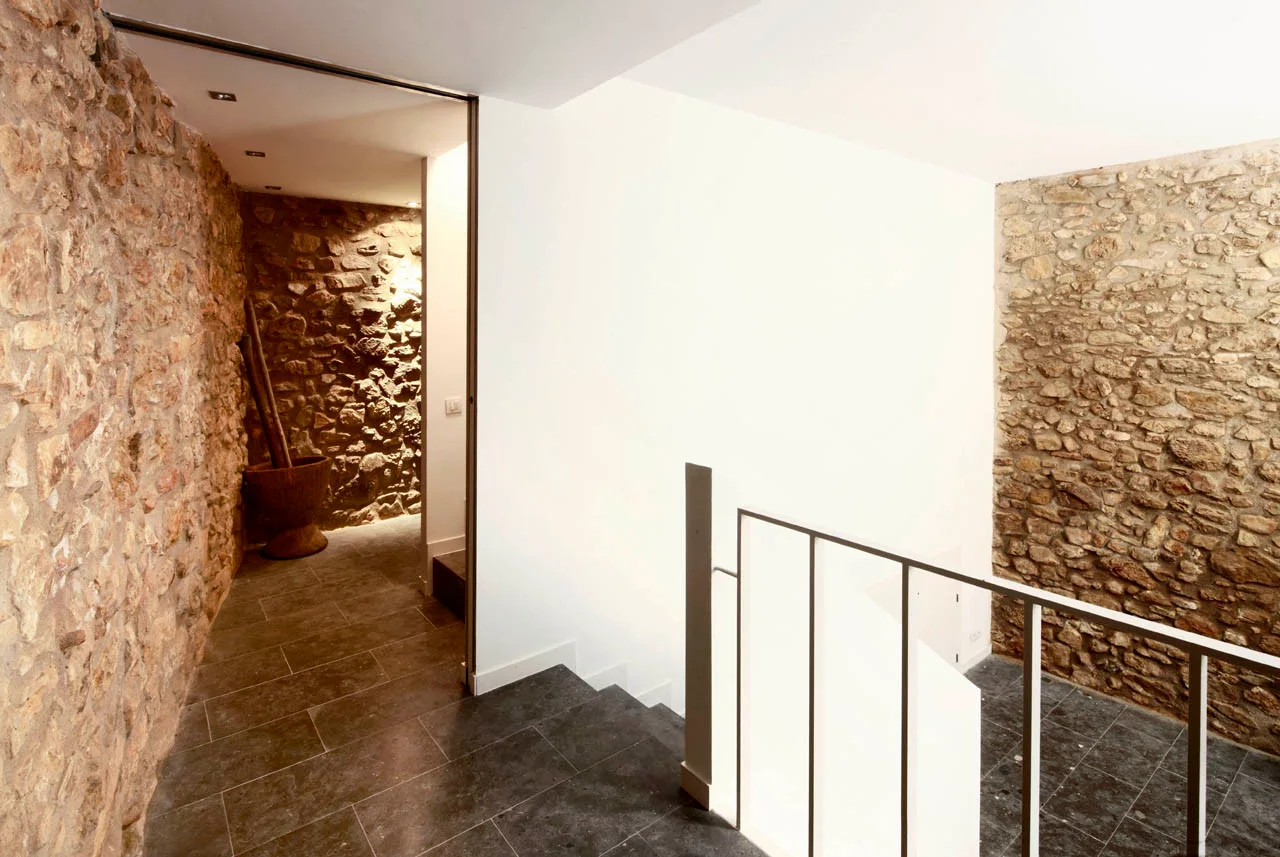 A modern staircase features dark stone flooring, minimalist metal railings, and contrasting rough exposed stone and smooth white walls.