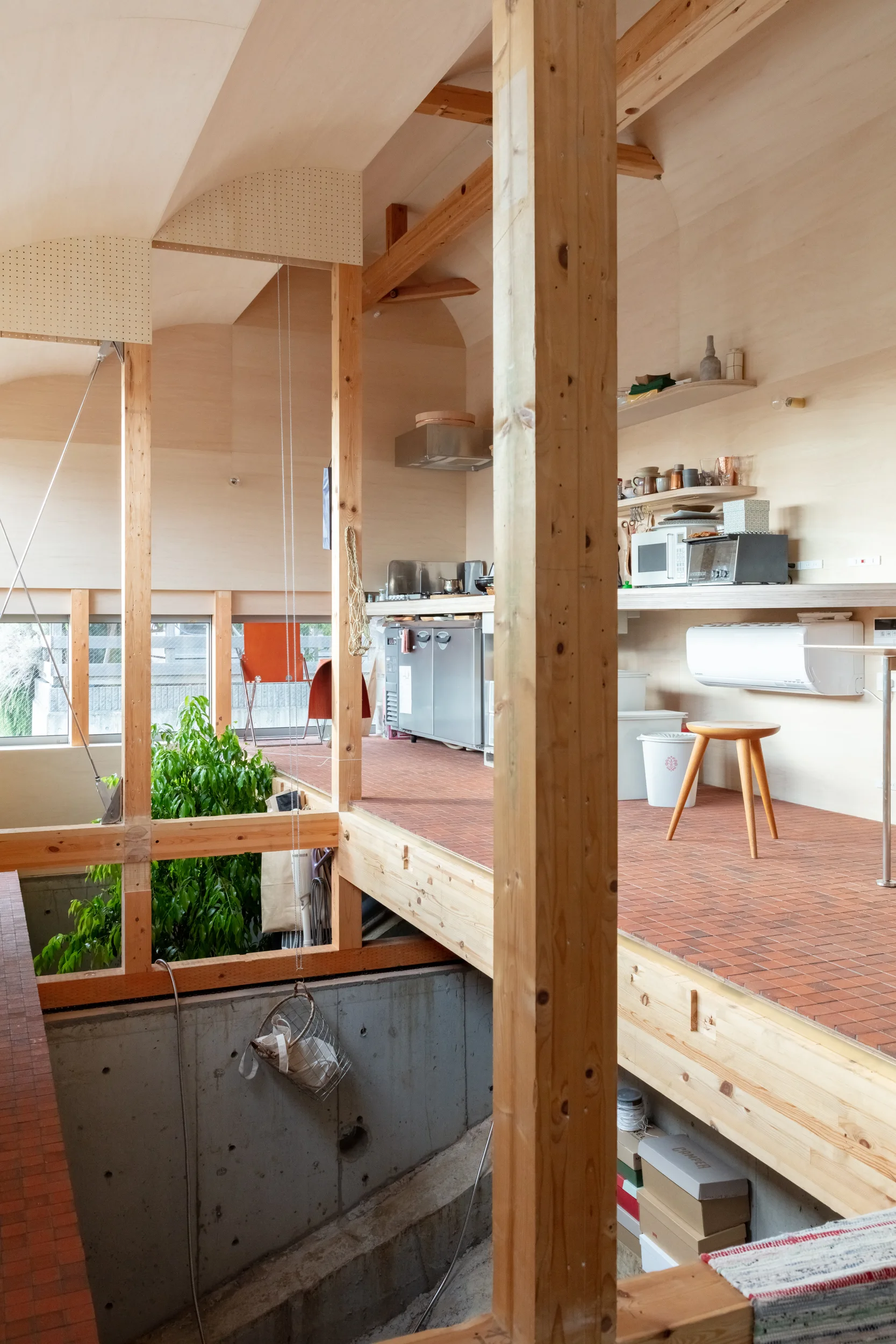 A split-level kitchen features plywood walls, exposed wooden beams, small terracotta floor tiles, and a cutout looking down to a concrete lower level.