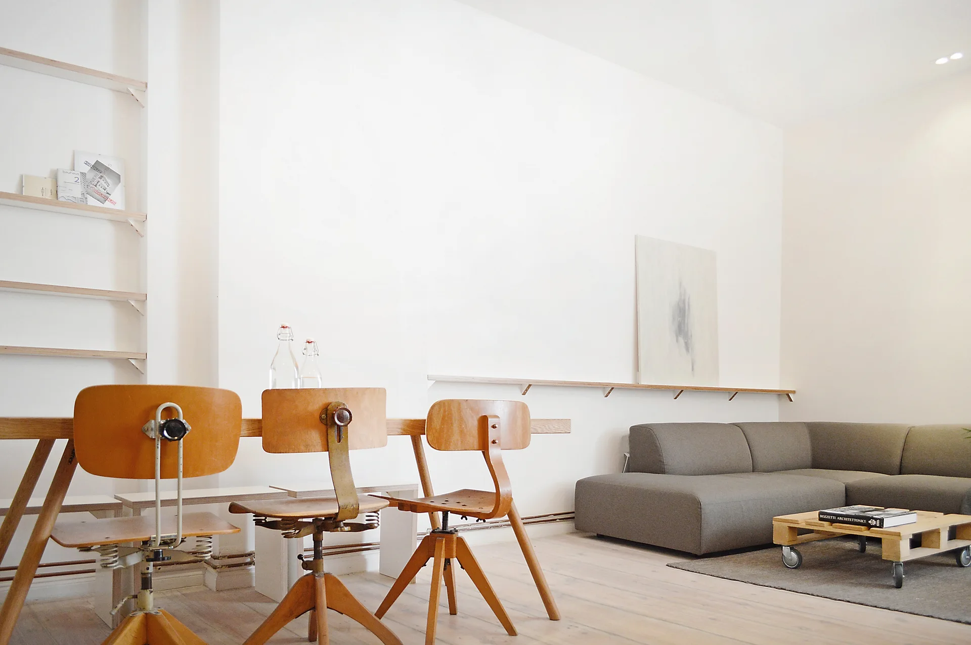 A minimalist living and dining space features three vintage wooden swivel chairs at a table, a low-profile grey sectional sofa, and a pallet coffee table.