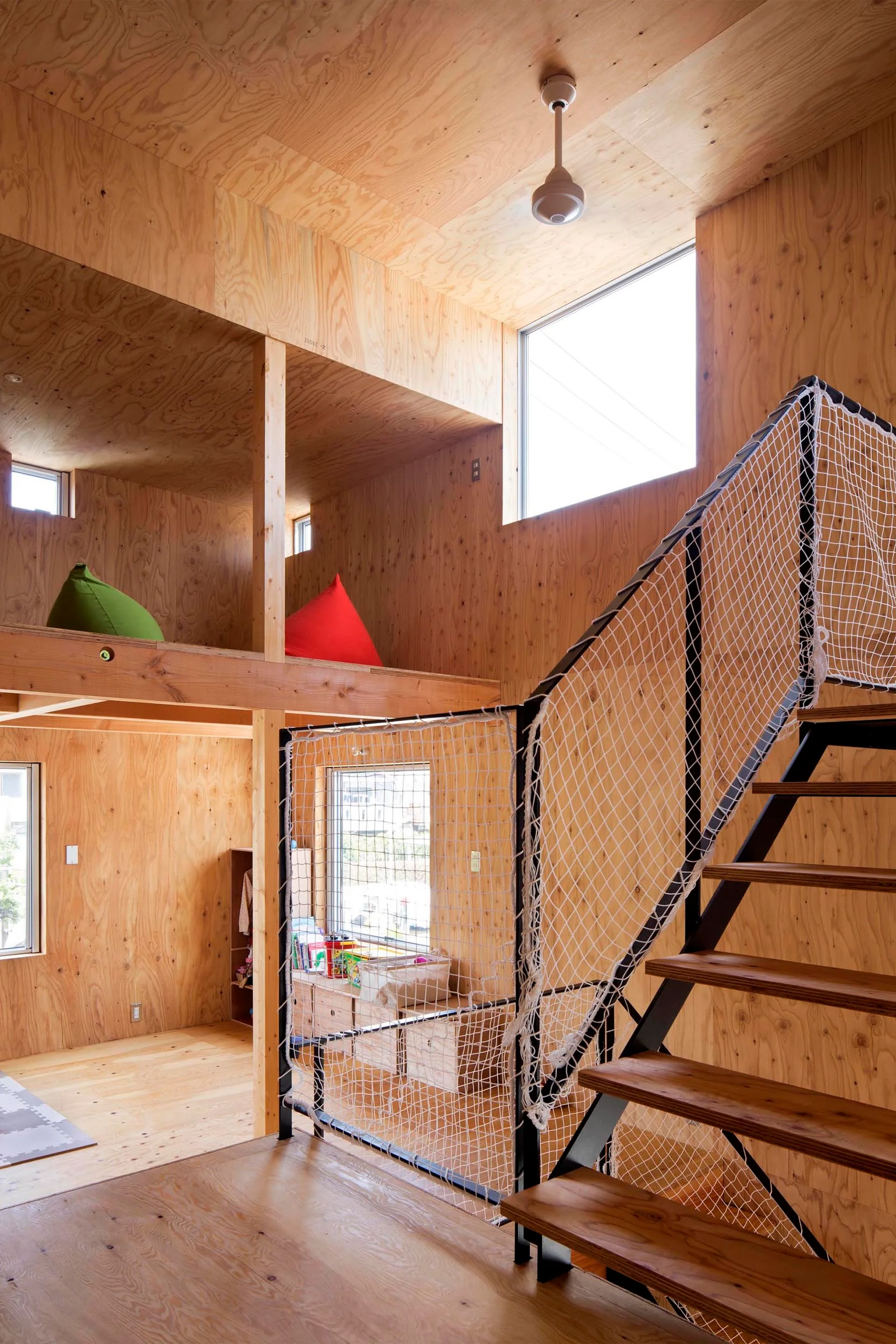 A modern loft interior completely clad in light-toned plywood, featuring an open-tread staircase with a white safety net railing.