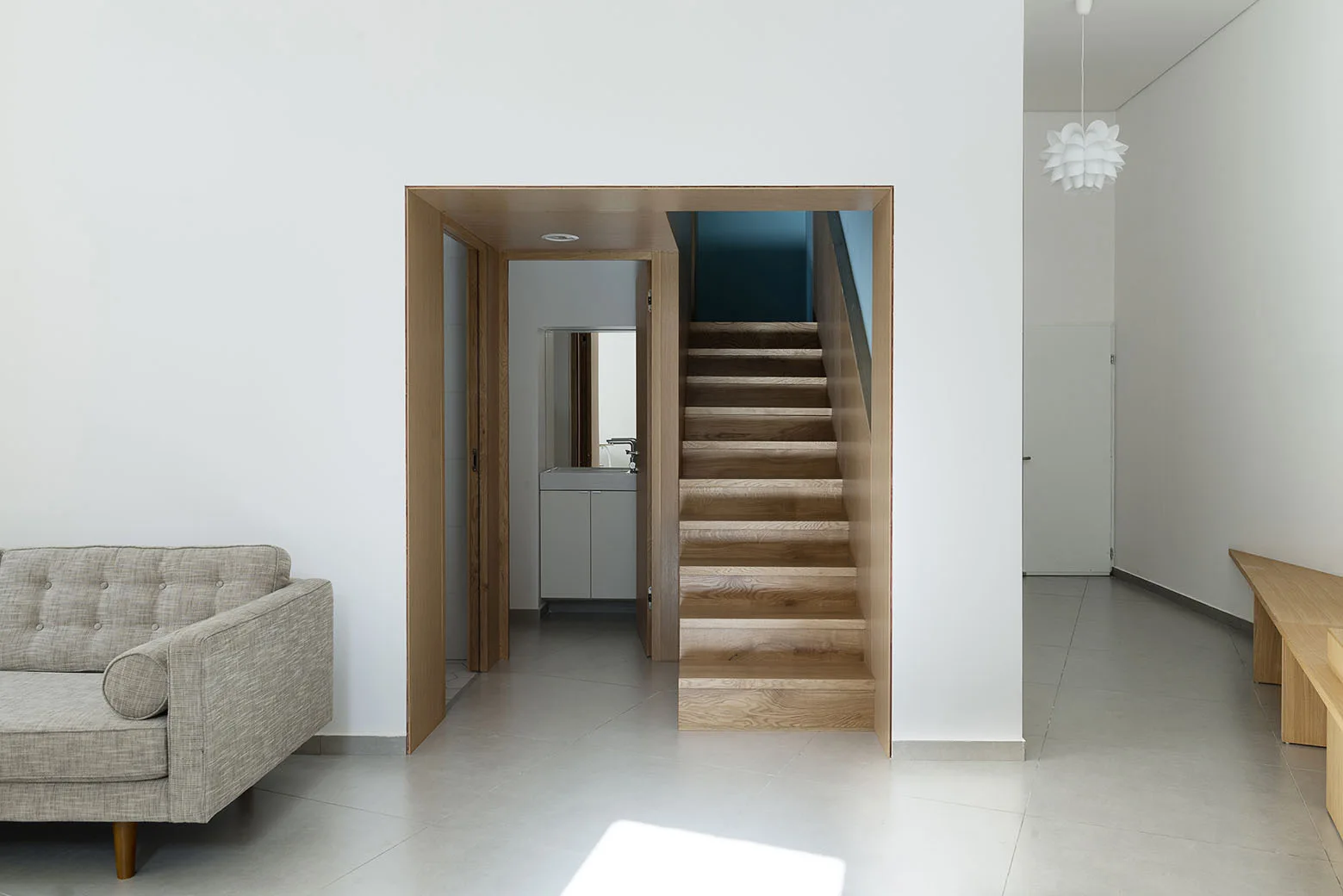 A wooden staircase framed by a wood-lined doorway in a white wall with a grey tiled floor and a grey sofa visible on the left.