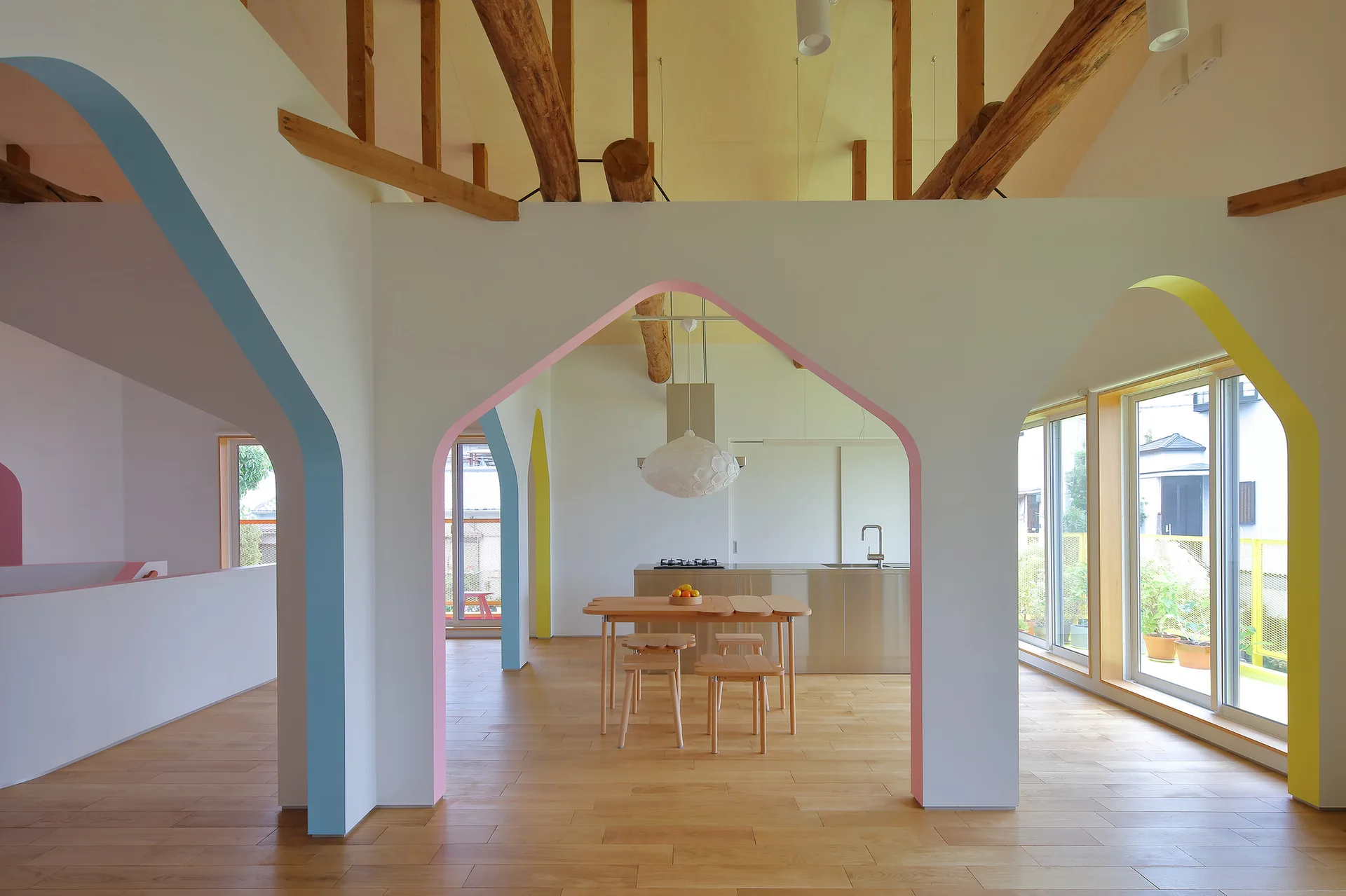 A bright open-plan kitchen and dining area features white partition walls with pastel-painted archways, exposed wooden ceiling beams, and a wood floor.