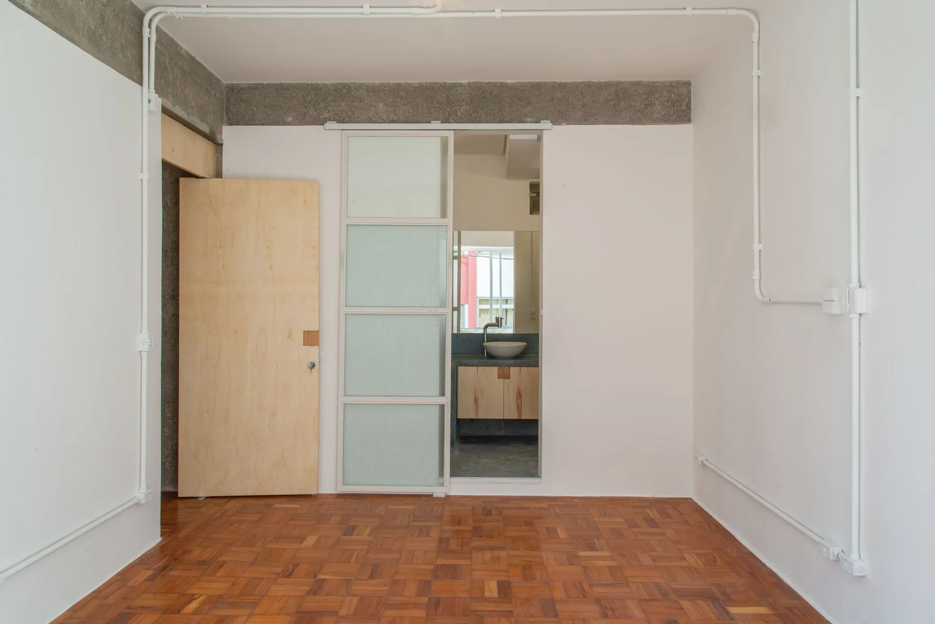 A minimalist room with warm wood parquet flooring, white walls, exposed conduit, and a framed sliding glass door leading to a bathroom.