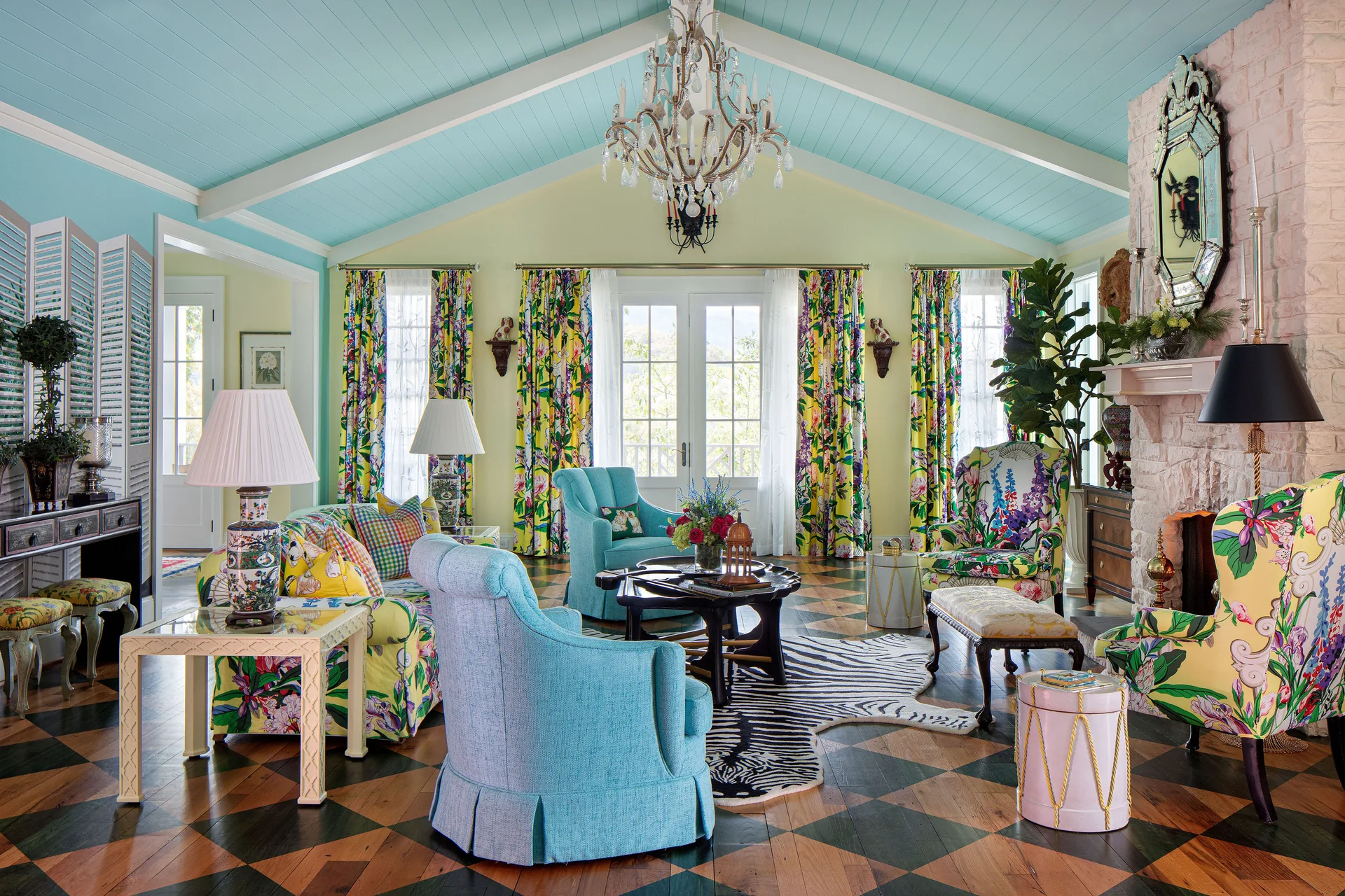 An eclectic living room features a blue vaulted ceiling, floral patterned sofas, a stone fireplace, and a checkerboard wood floor.