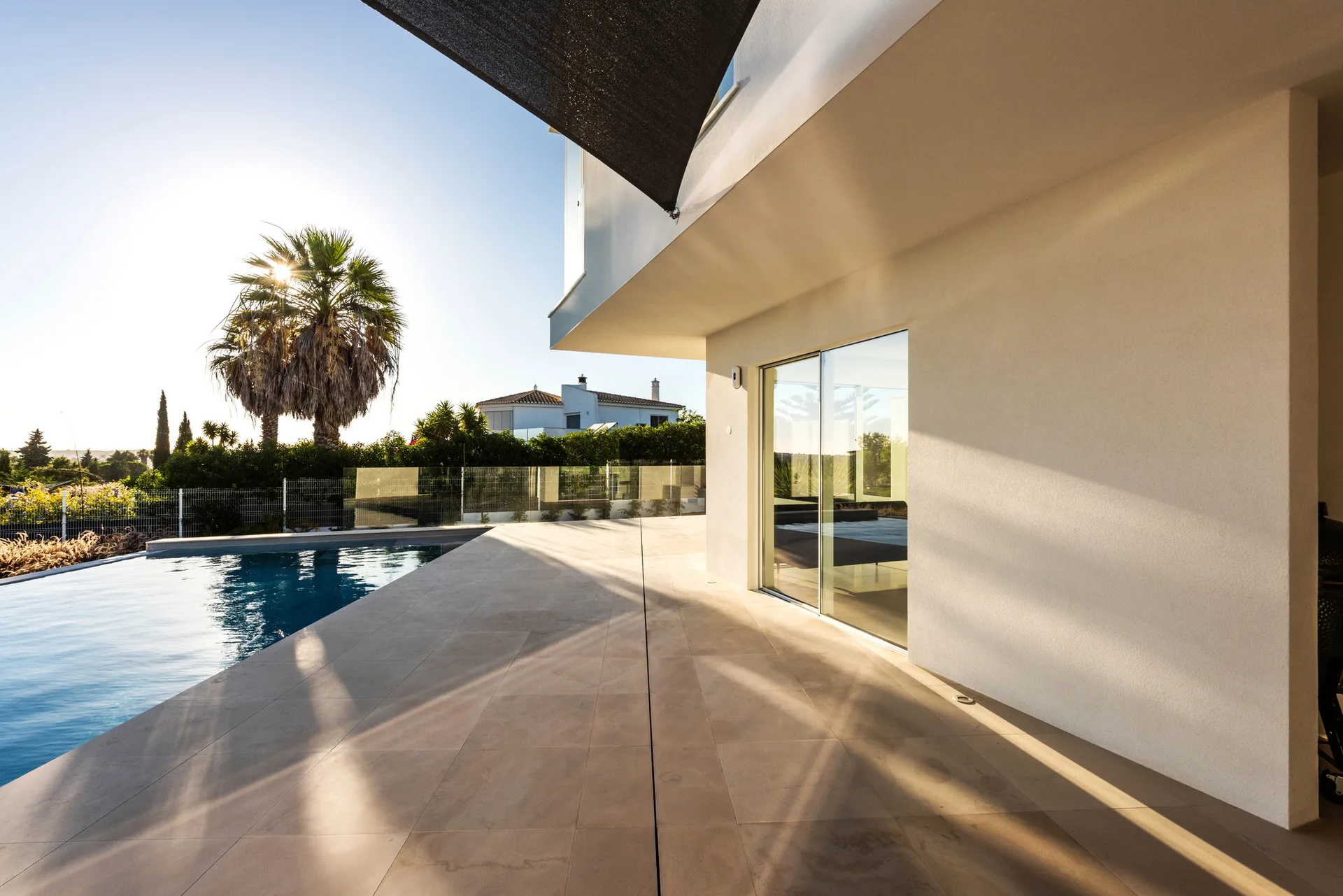 A modern residential patio features large-format beige tiles alongside a blue swimming pool under a bright sky with a dark sun shade overhead.