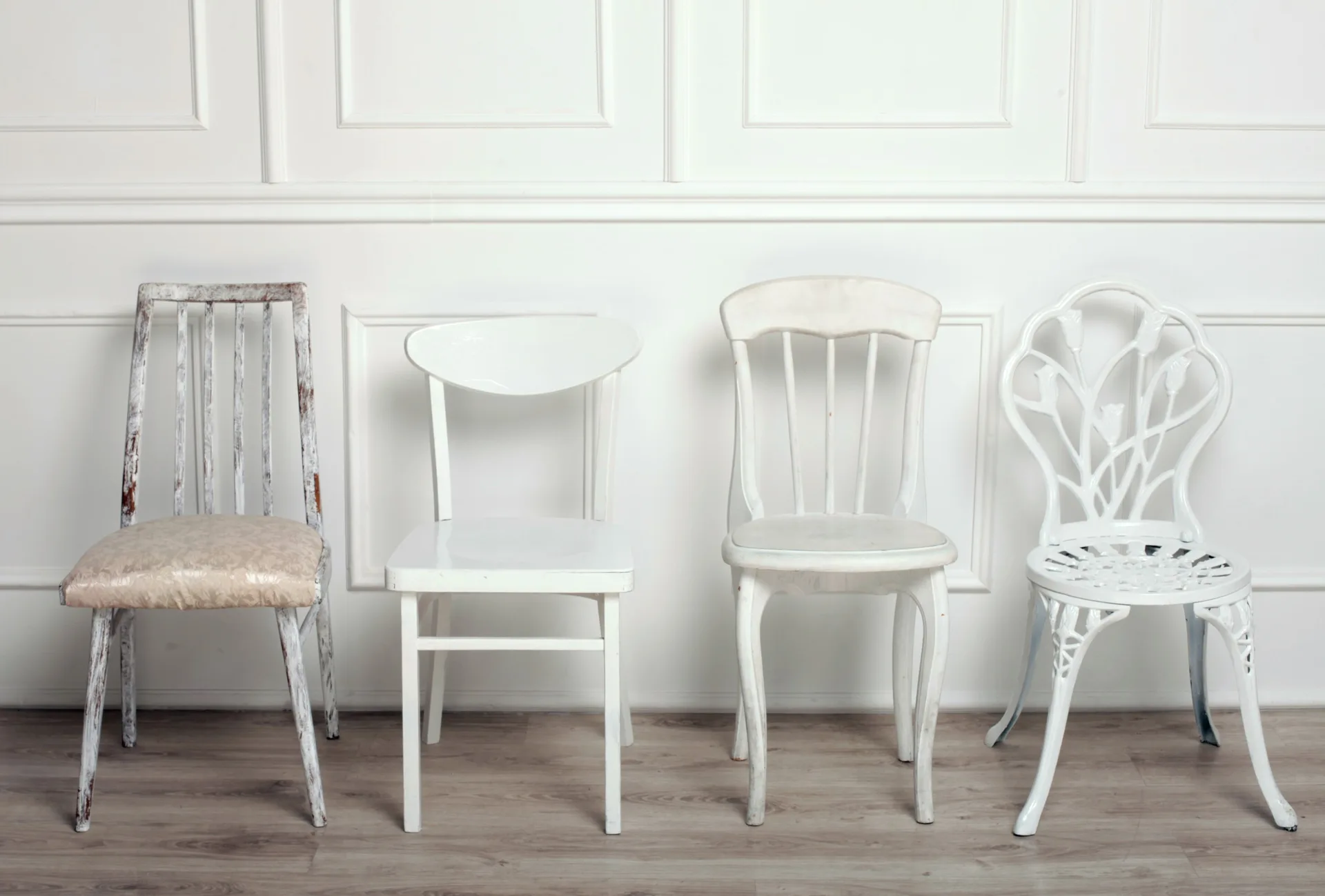 Four mismatched white and off-white chairs in different styles are lined up against a white paneled wall with classic molding on a light wood floor.