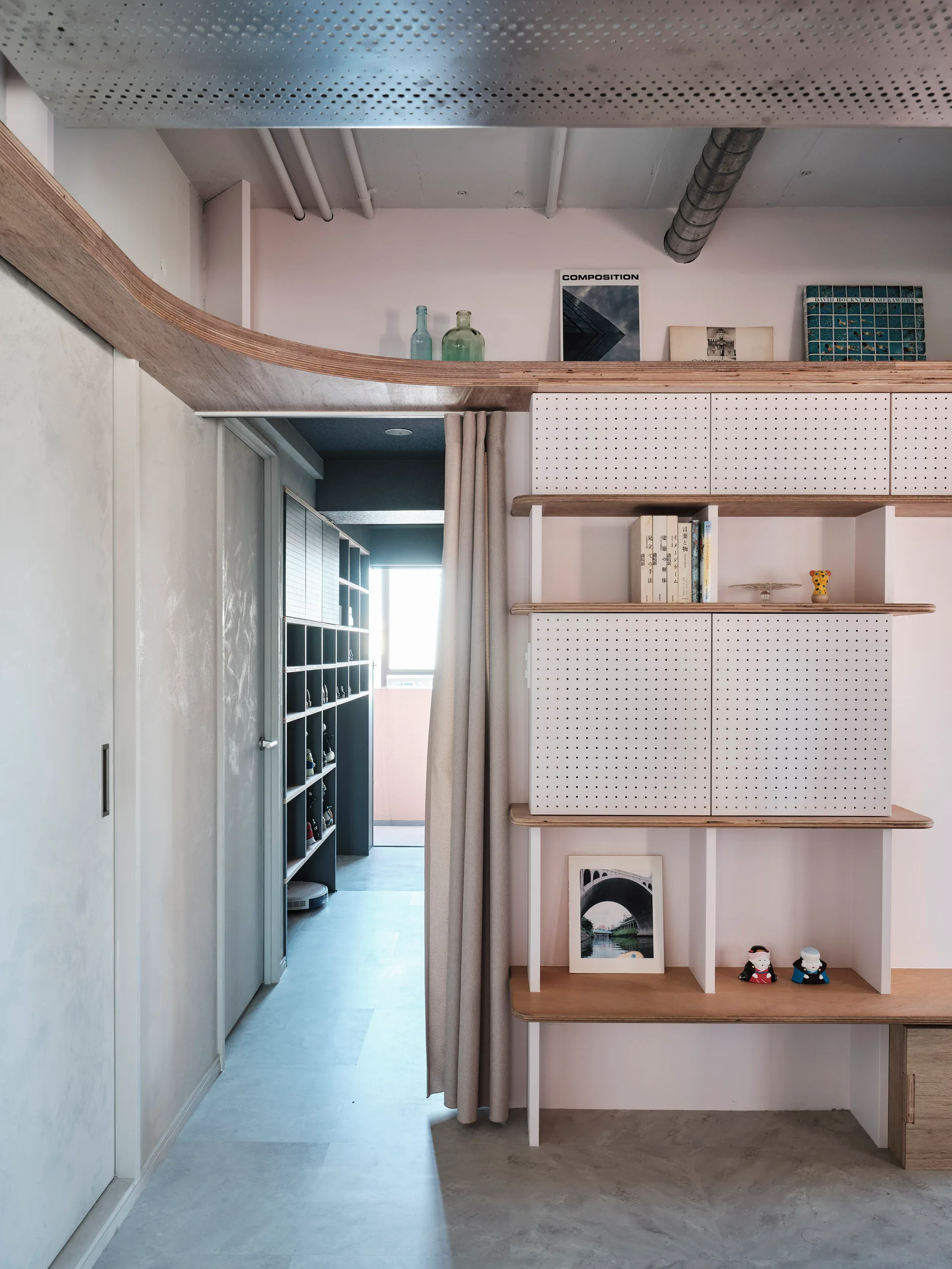 A minimalist hallway features a custom white pegboard shelving unit, light pink walls, curved plywood trim, and a beige fabric curtain.