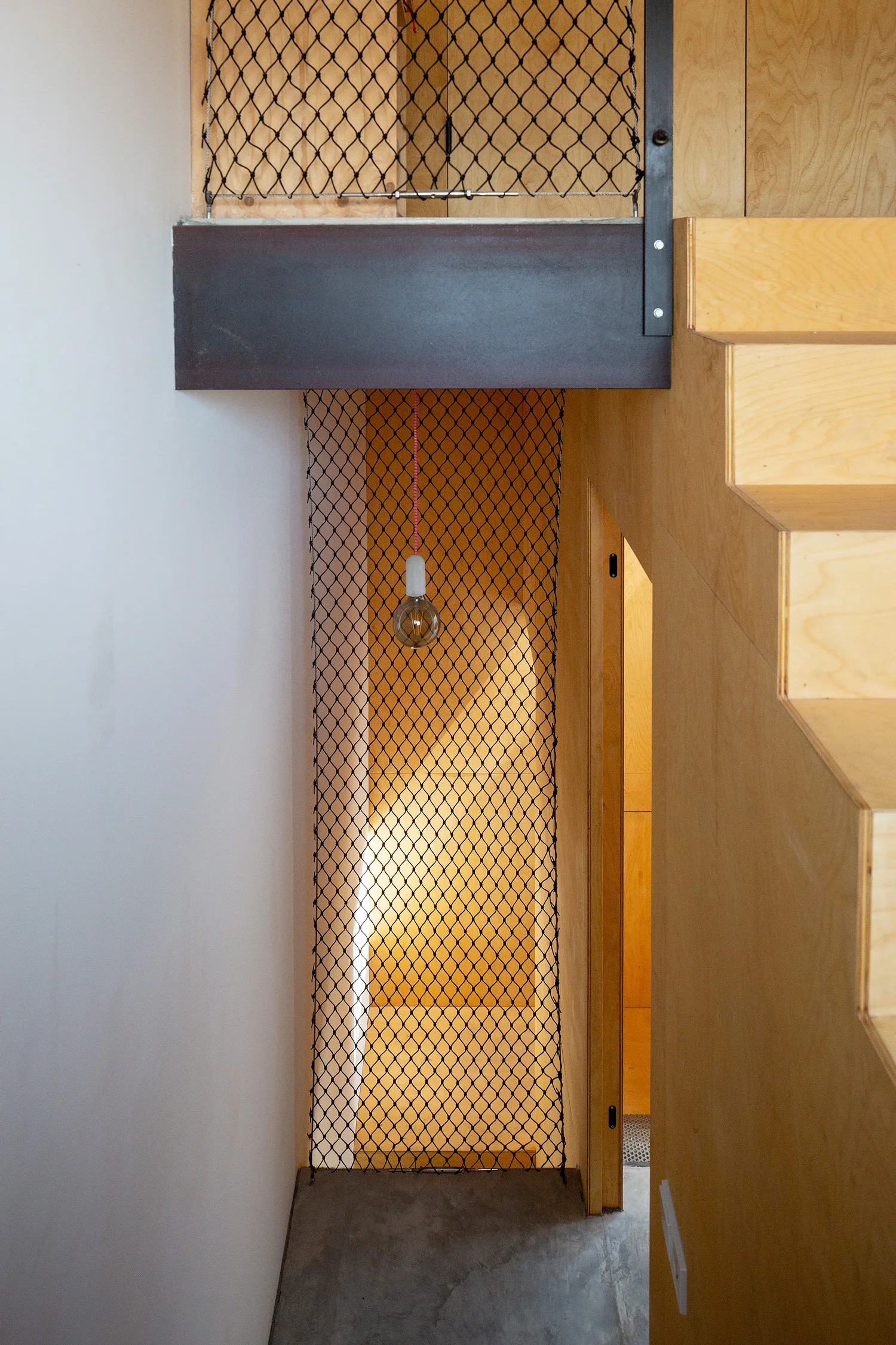 A modern plywood staircase features black mesh safety netting as a banister and a single minimalist hanging pendant light.