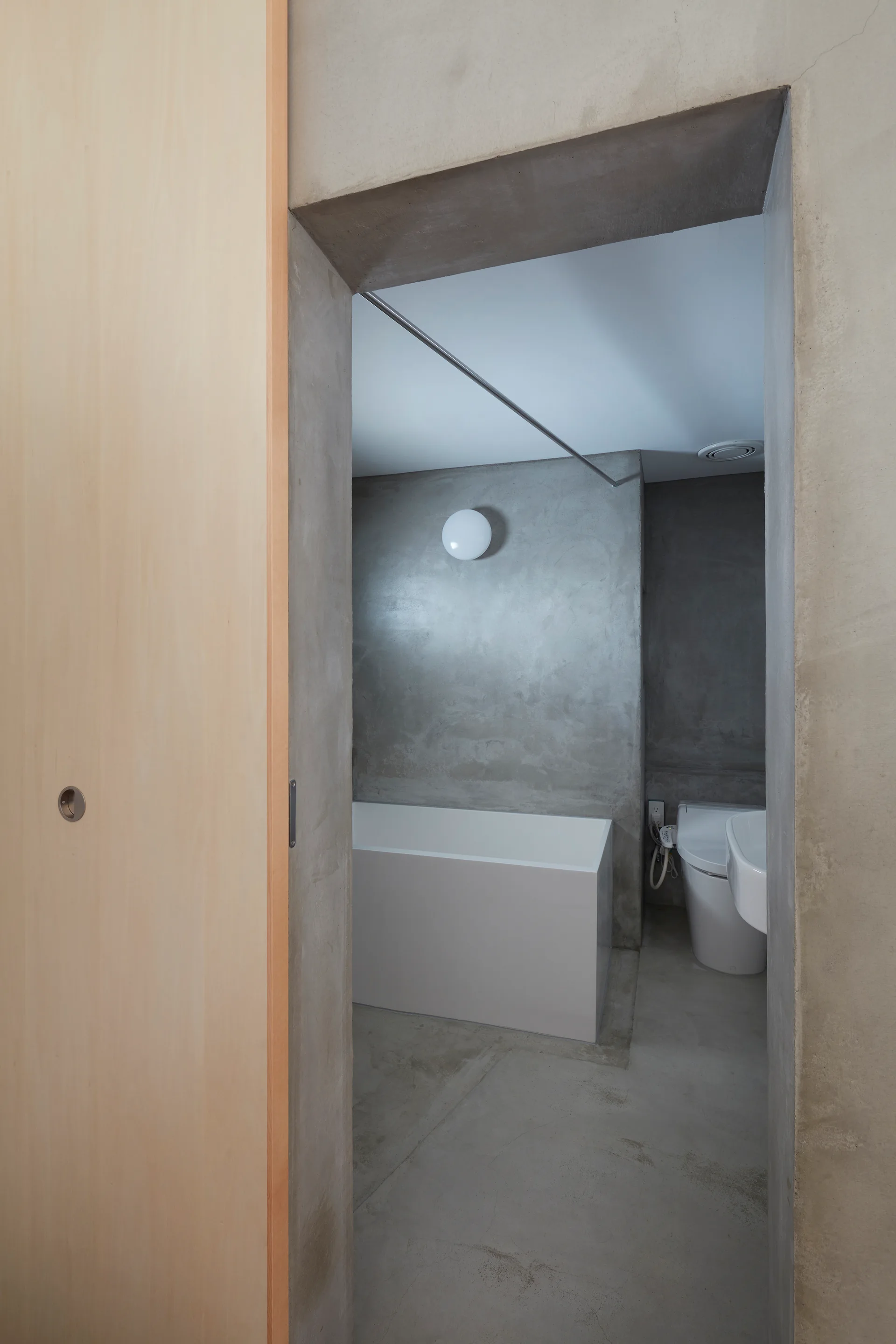 A minimalist bathroom view featuring smooth concrete walls, a white rectangular bathtub, a white toilet, and a light wood sliding door at the entrance.