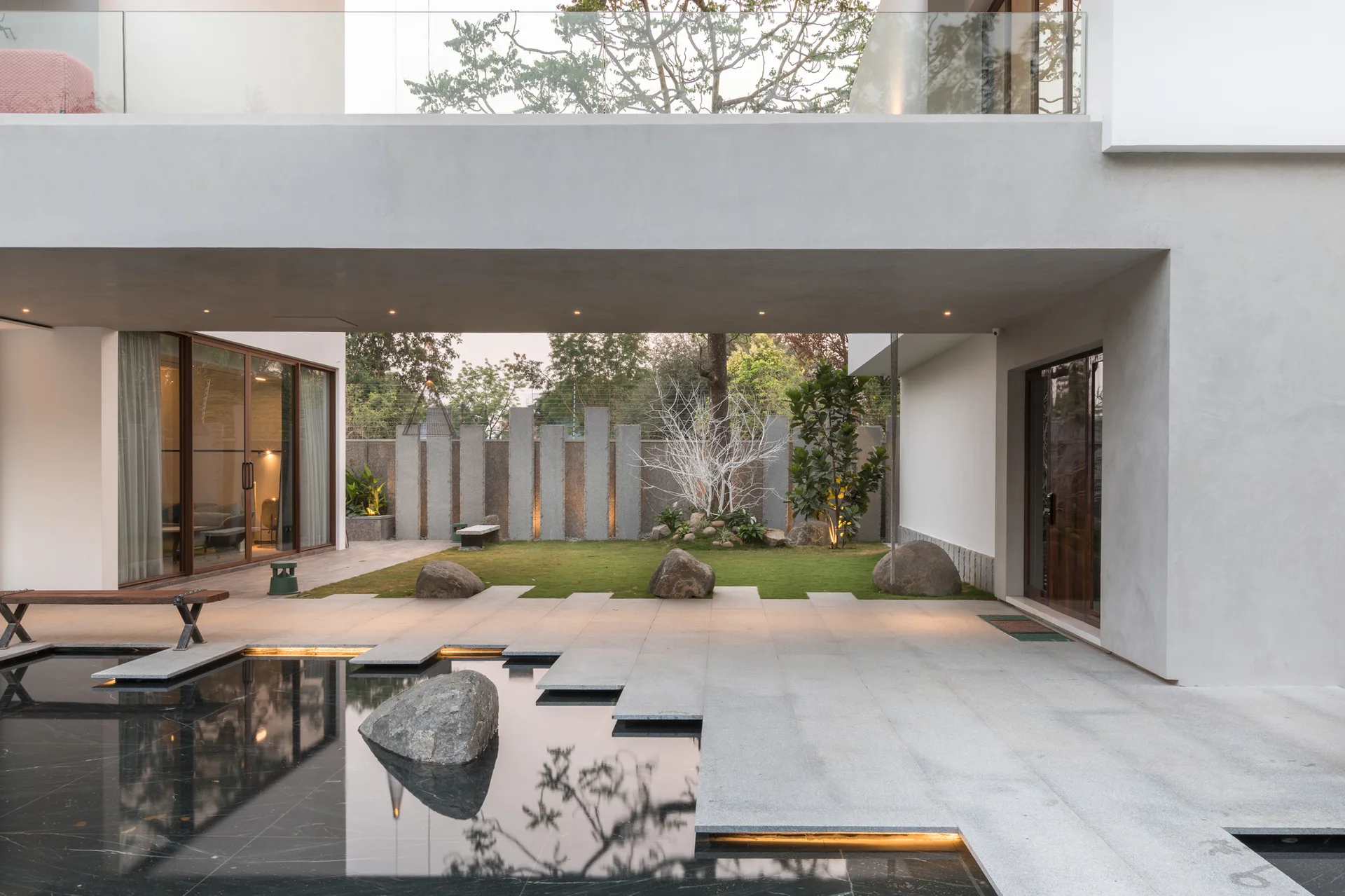 A modern residential courtyard features a stone-paved patio, a black sunken water feature with a large boulder, and a grassy lawn under a concrete overhang.