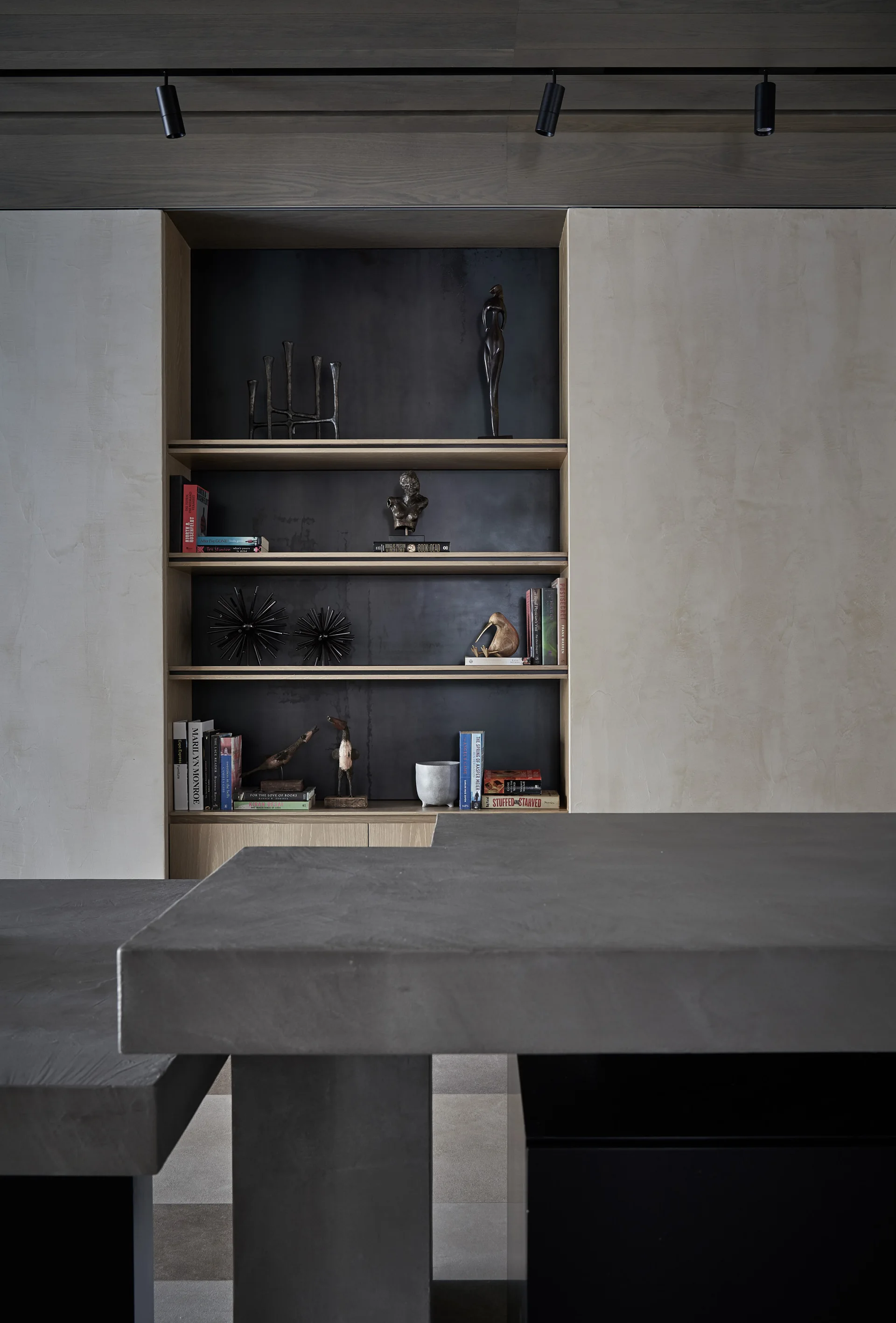 A built-in shelving unit with dark metal back panels and wood trim stands behind a gray concrete countertop and plaster walls.