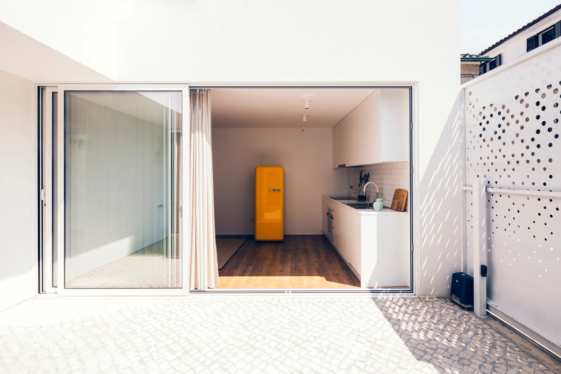 A minimalist white kitchen with a bright yellow retro refrigerator is visible through wide sliding glass doors opening onto a sunny stone patio.