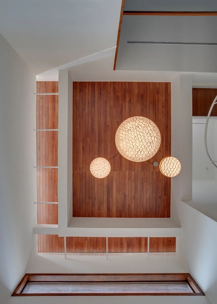 A low-angle view looking up at a slatted wood ceiling framed by white beams, featuring three glowing circular pendant lights of varying sizes.