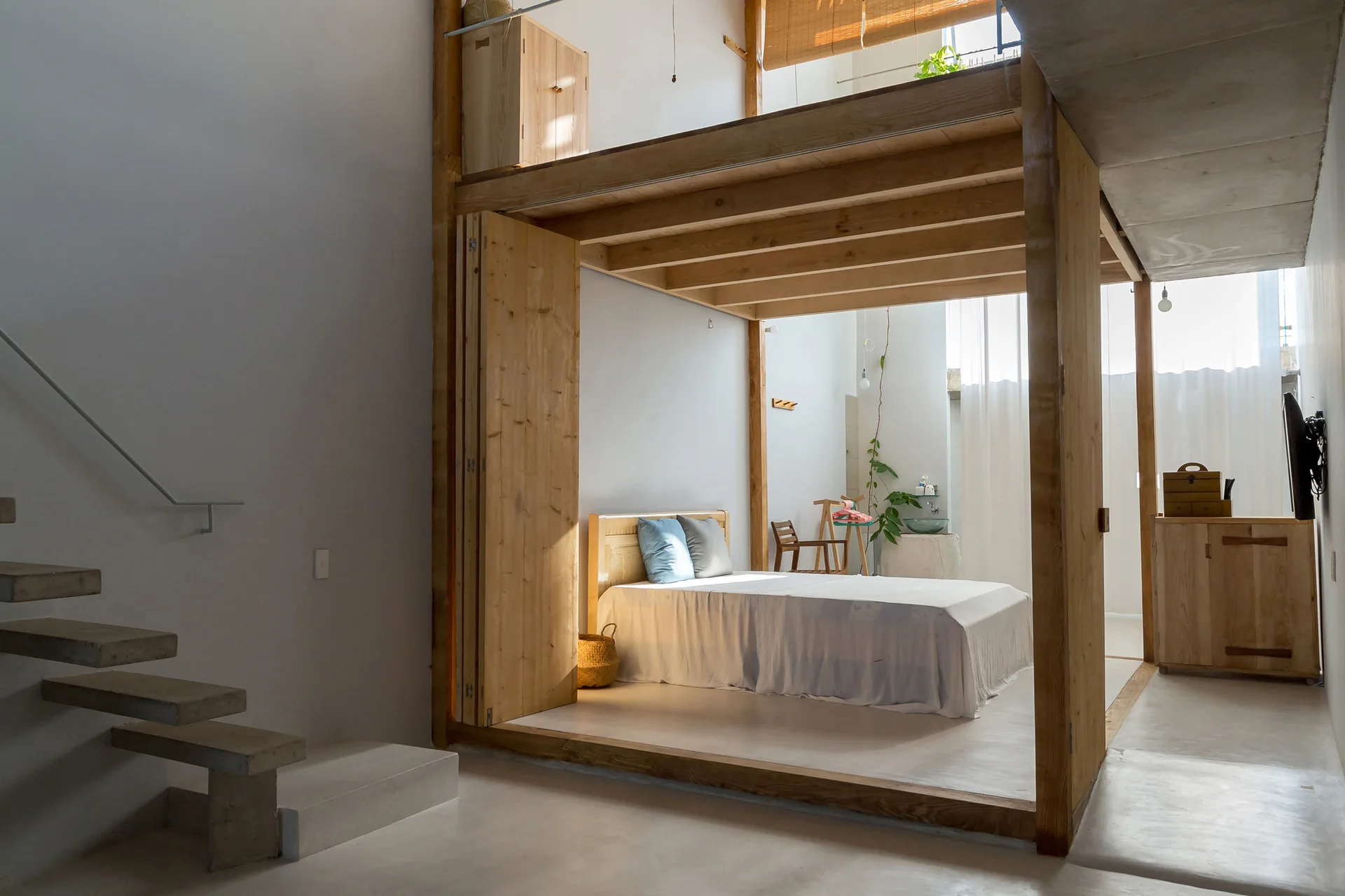 A minimalist bedroom features a bed framed by a light wood post-and-beam structure beneath a mezzanine, next to floating concrete stairs.