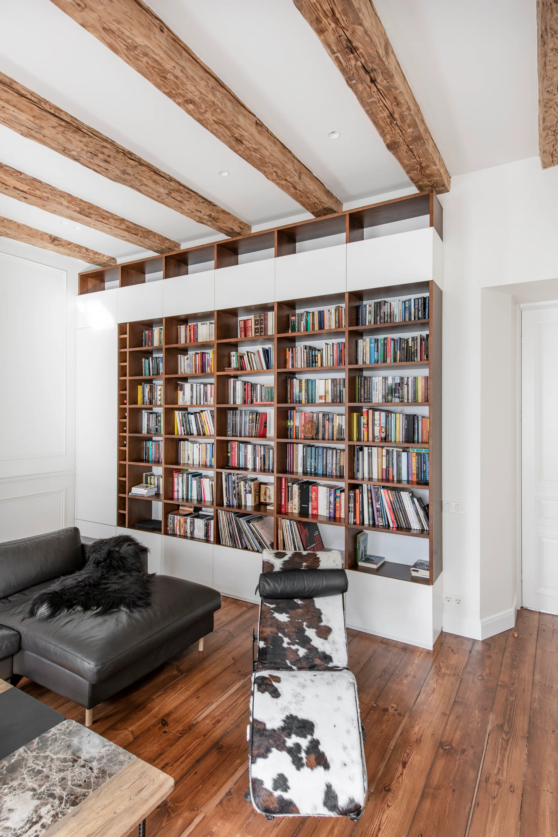 A living room featuring a floor-to-ceiling built-in wooden bookshelf, exposed ceiling beams, a black leather sofa, and a cowhide print lounge chair.