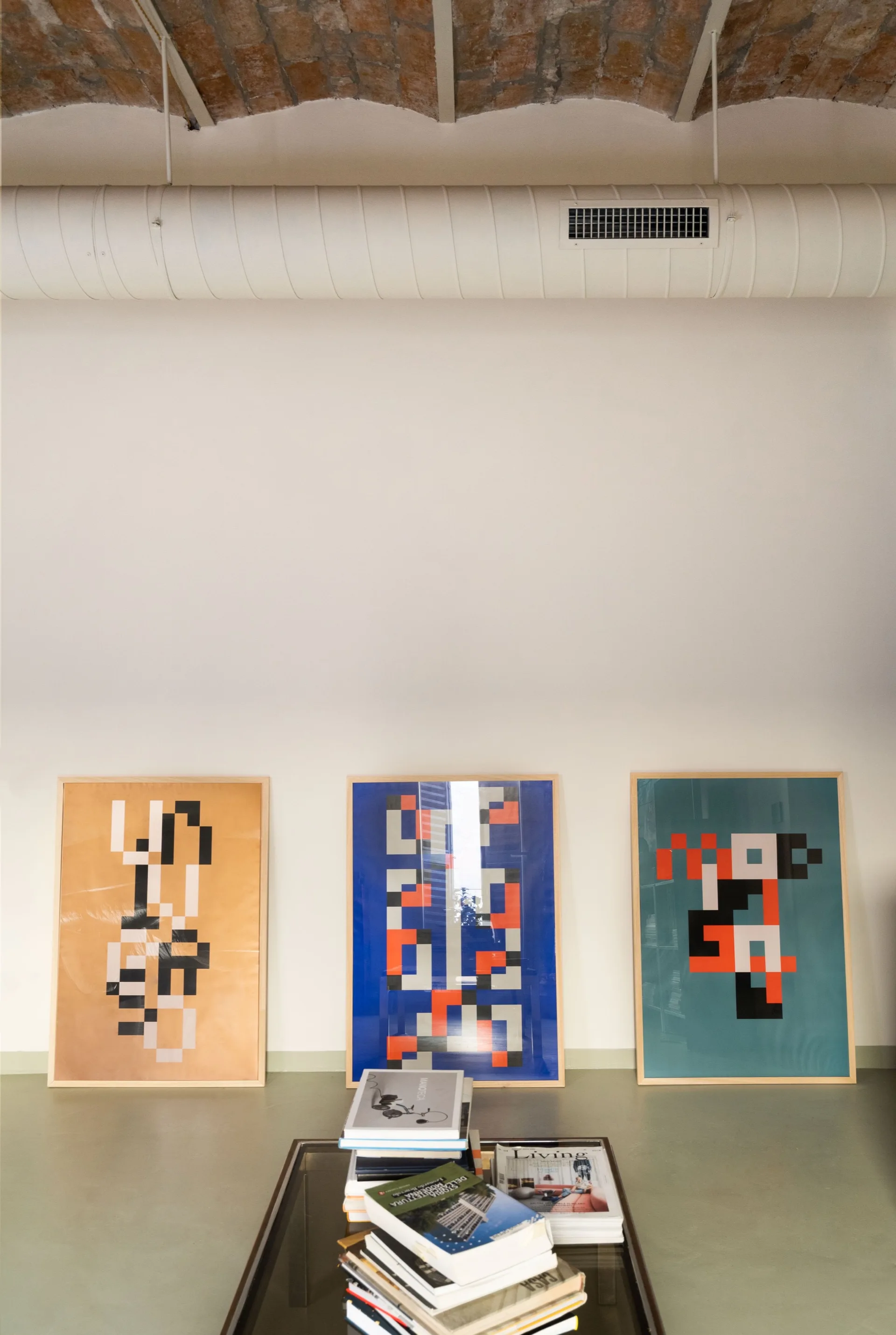 Three framed geometric posters lean against a white wall on a light green floor beneath an exposed brick vaulted ceiling.