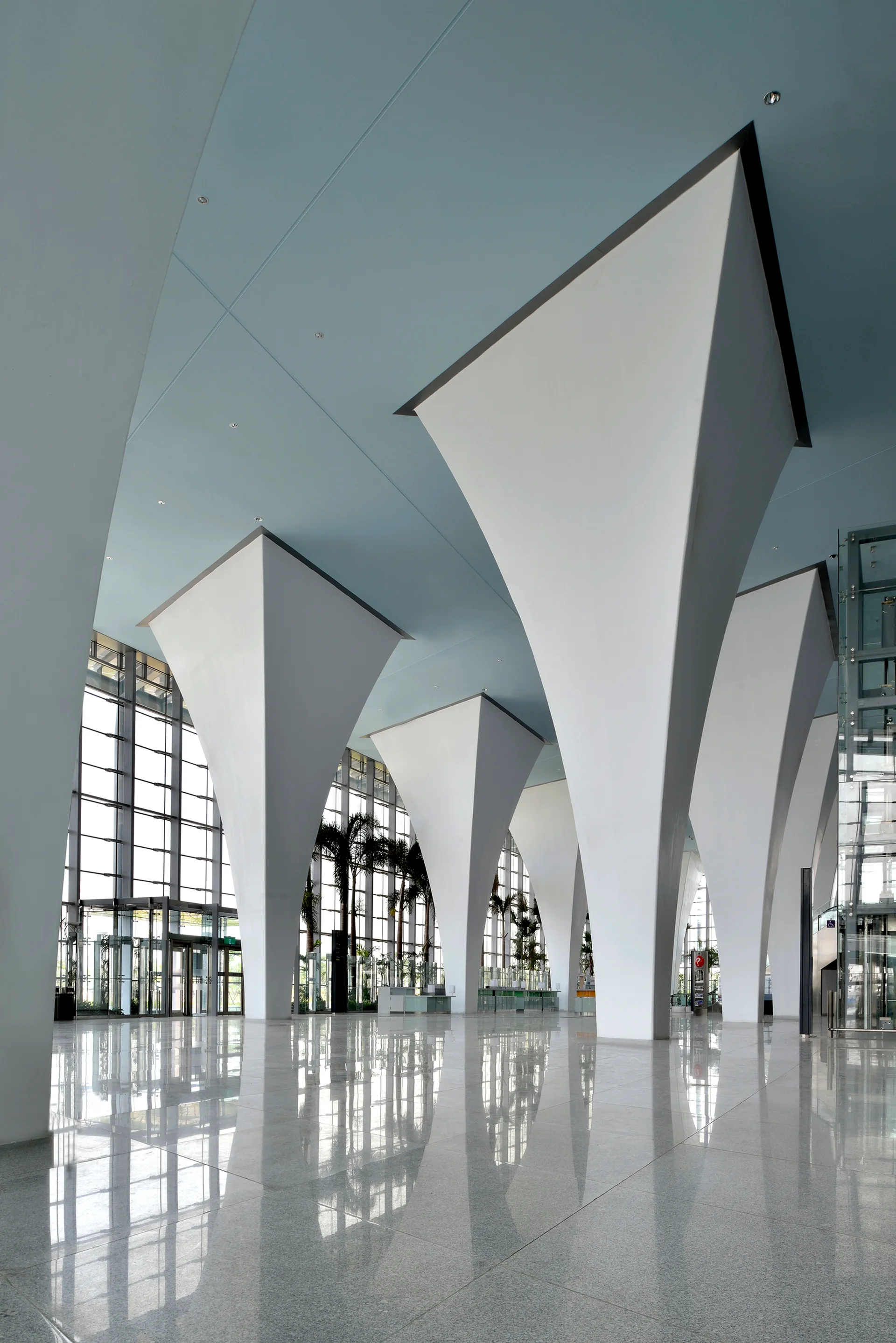 A spacious atrium with large, flared white columns supporting a light blue ceiling over a highly reflective polished stone floor.