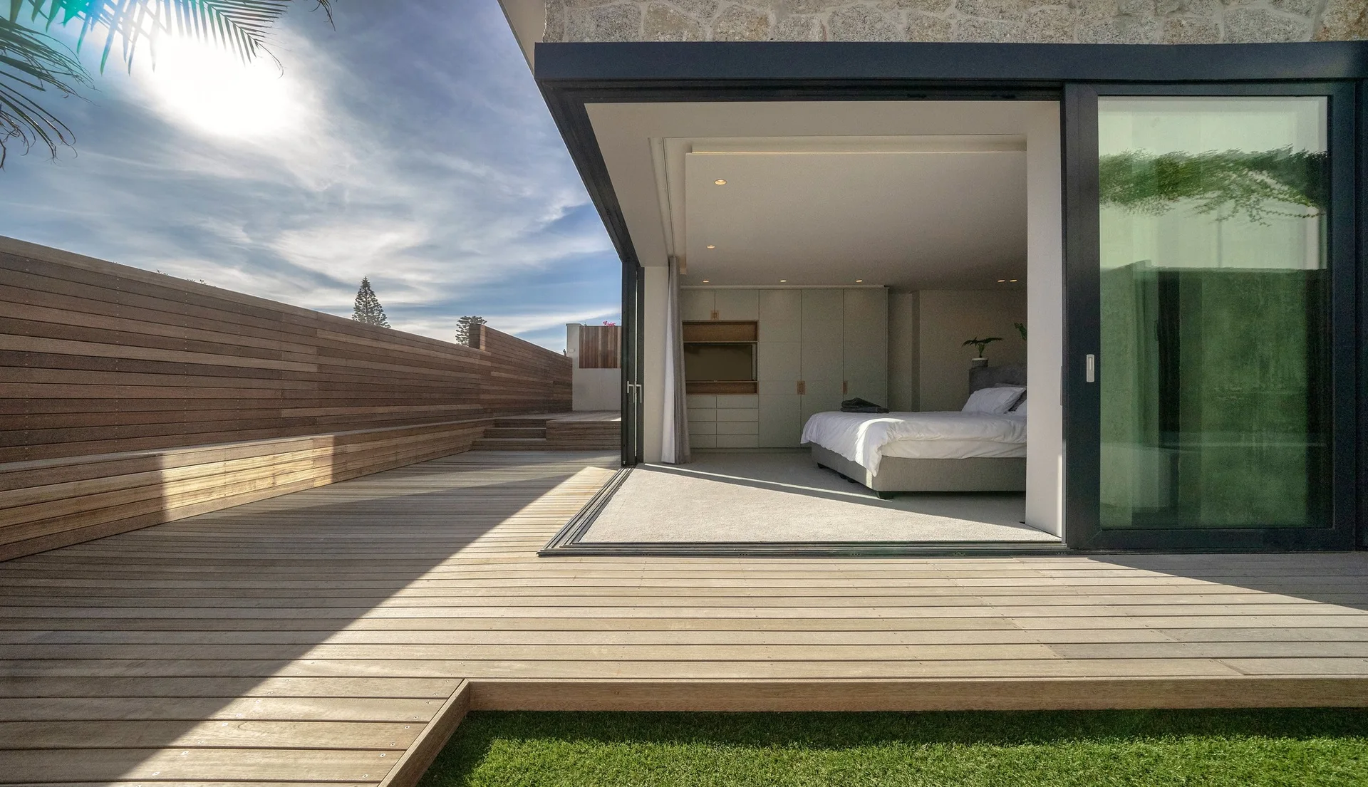 A modern bedroom with built-in cabinetry opens onto an expansive wooden plank deck with a high timber privacy fence.