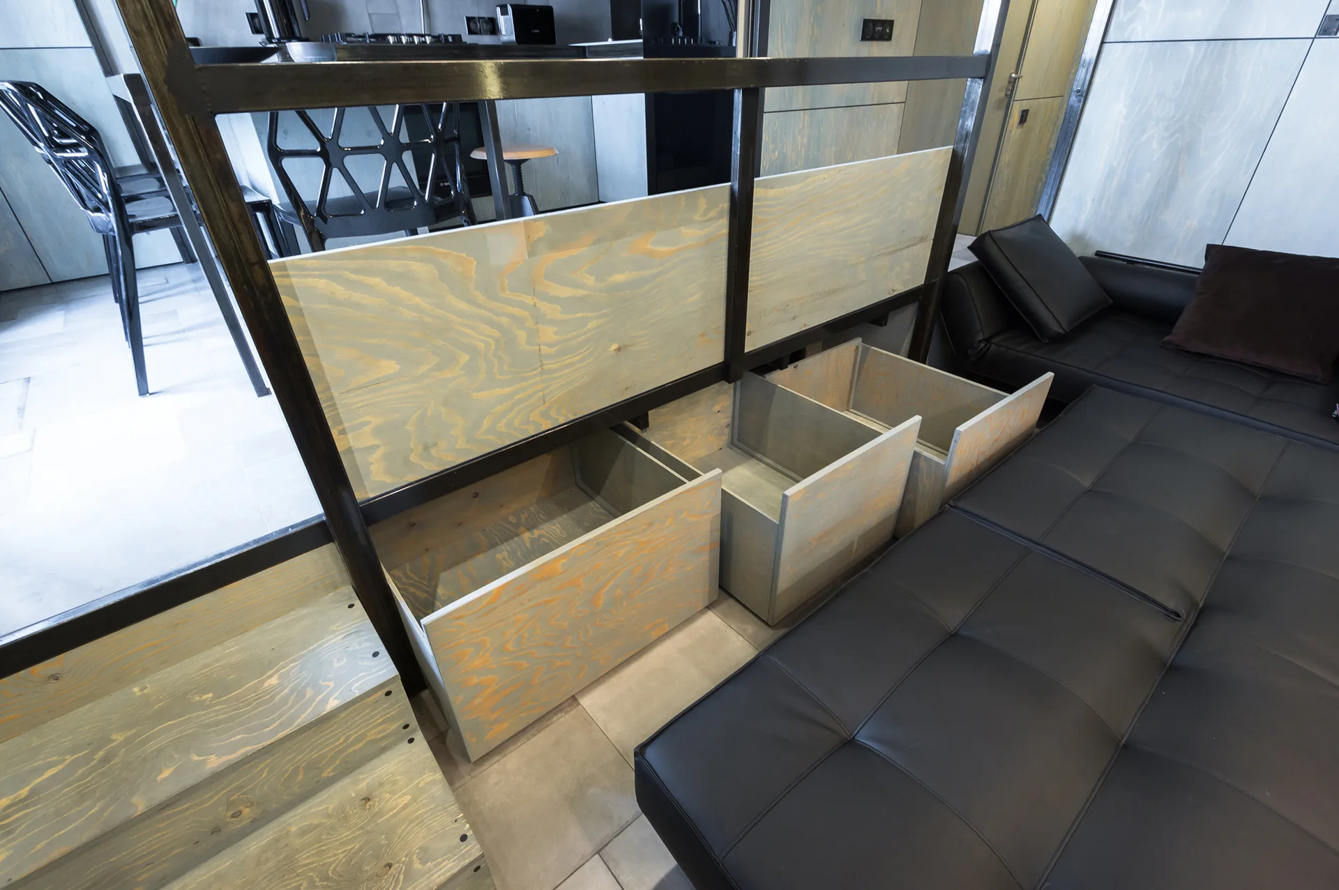 A modern living room features light plywood drawers pulled out from under a staircase next to a black leather sofa.