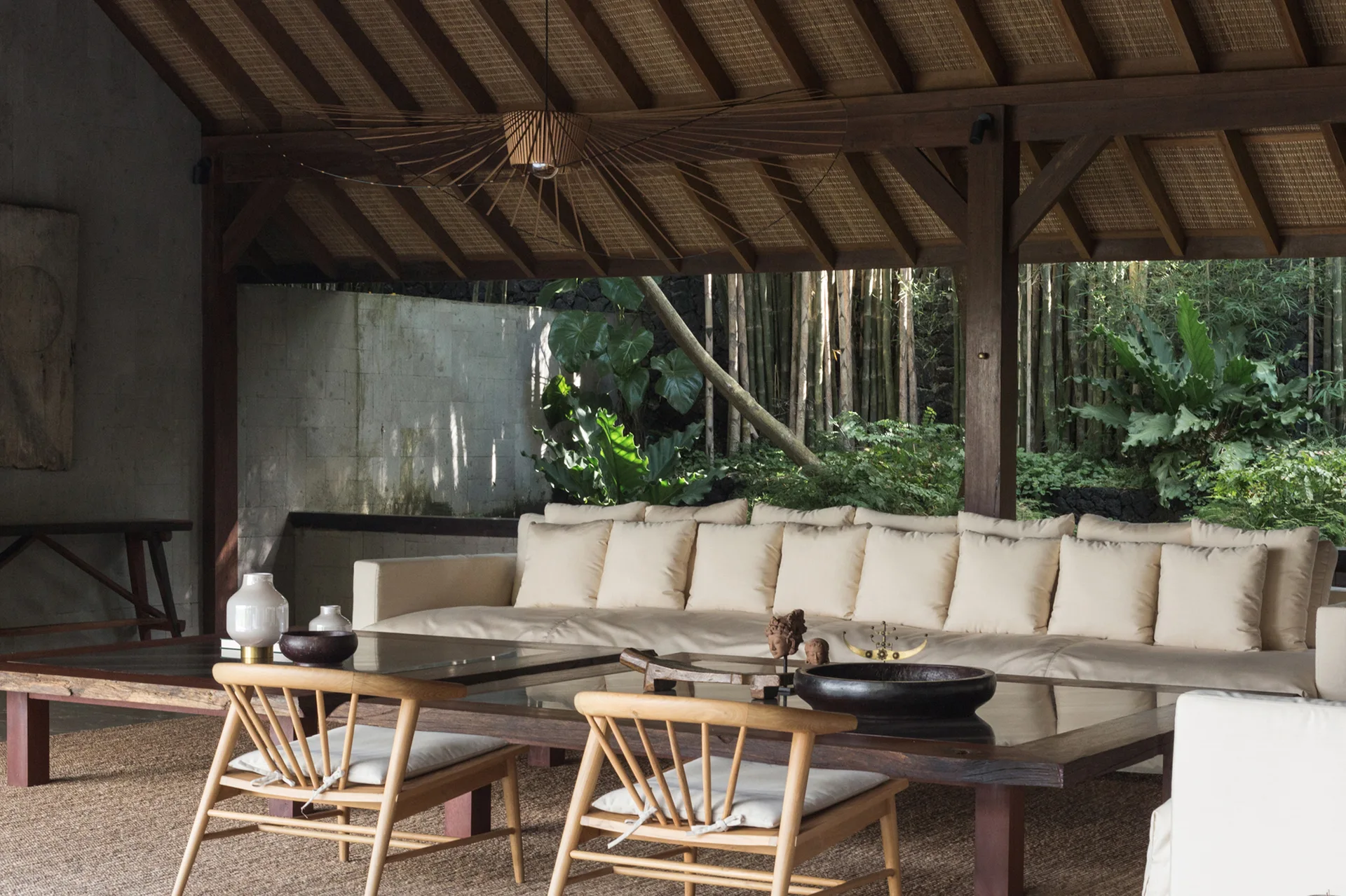 An open-air pavilion lounge features a long cream sofa and low wooden coffee table beneath a sloped thatched ceiling looking out to a garden.