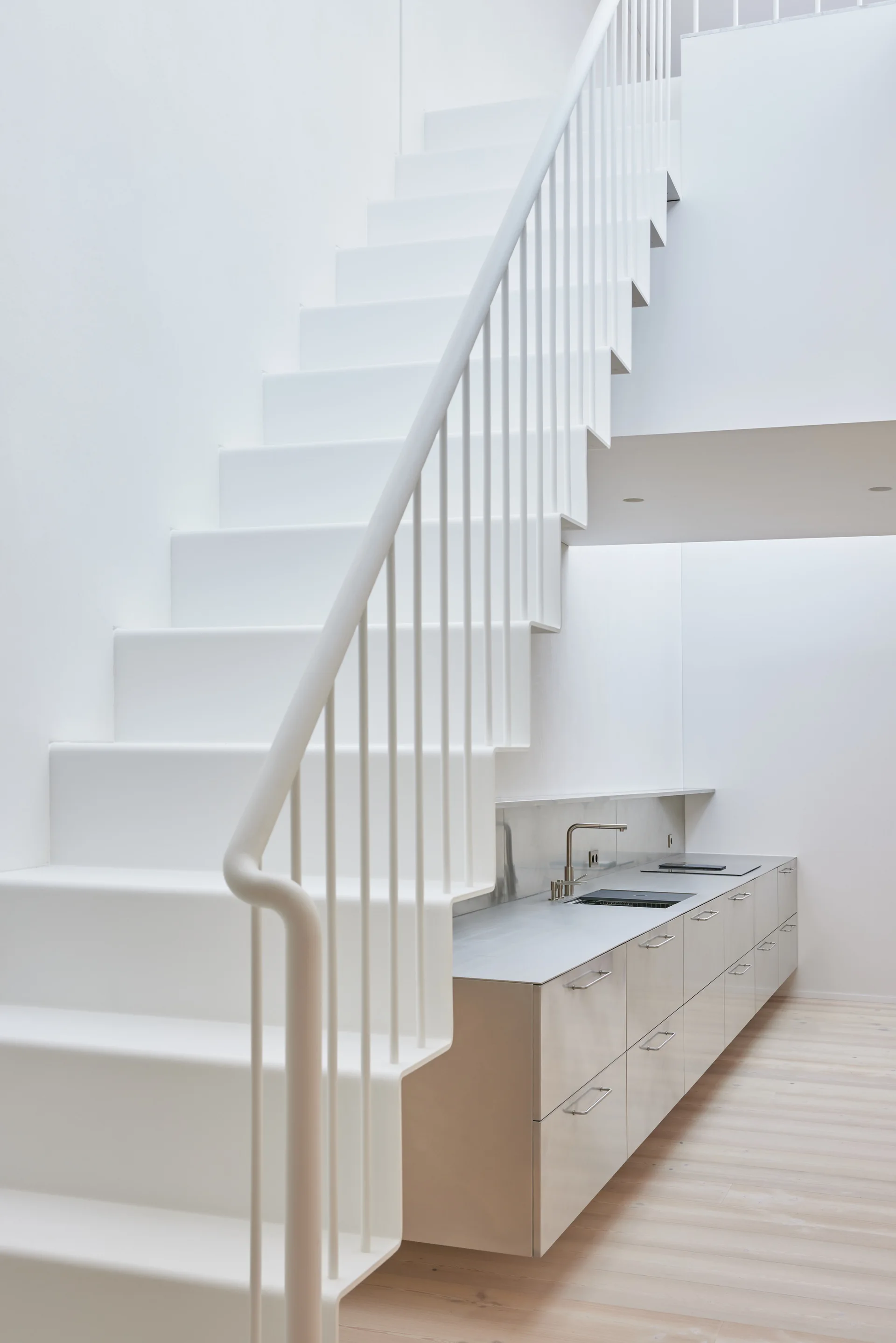 A minimalist white metal staircase ascends above a sleek grey kitchen cabinet unit with a sink and cooktop on light wood floors.