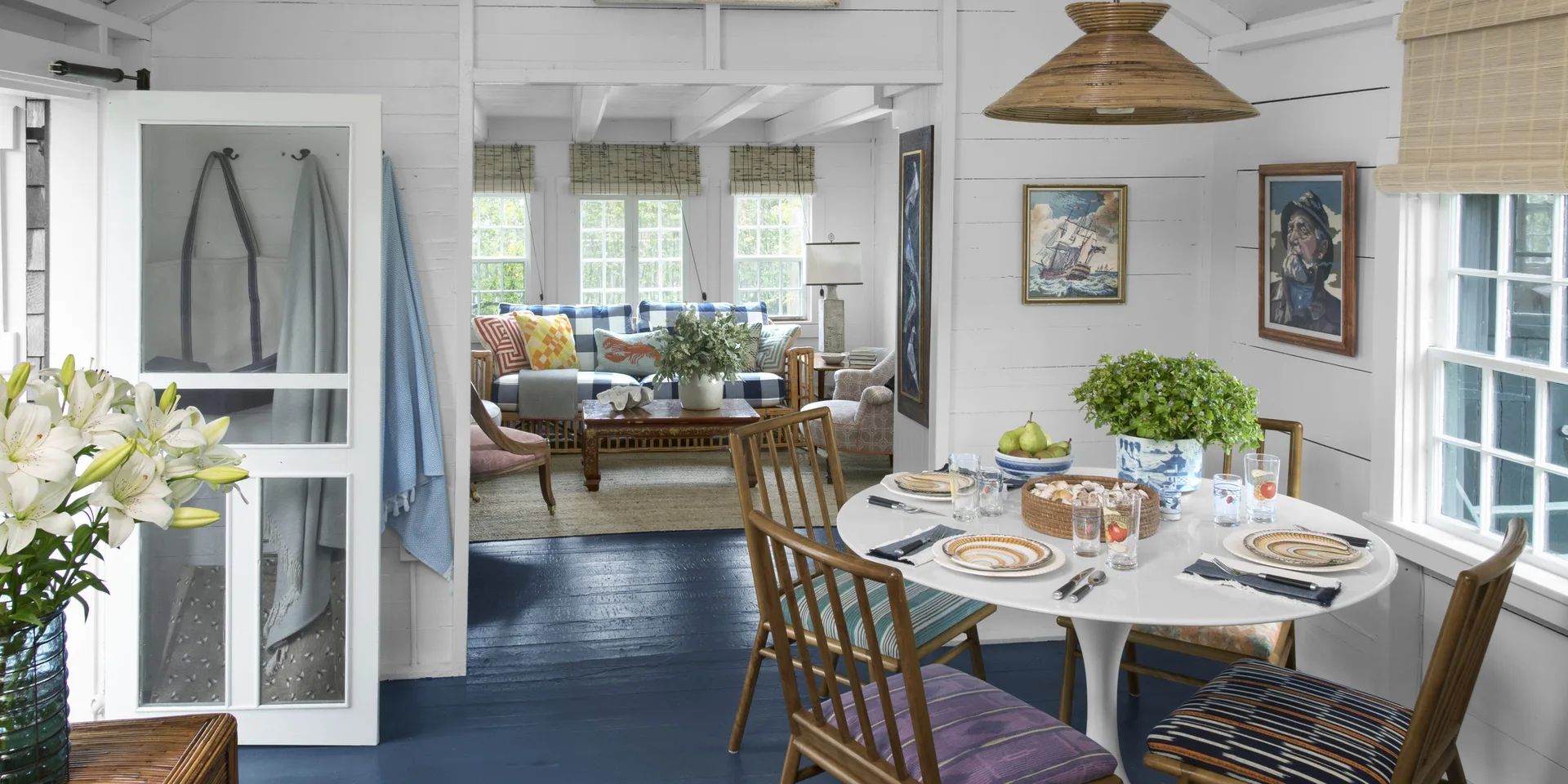 A cottage dining area featuring a round white table, wooden chairs, a woven pendant light, and a painted deep blue floor leading to a living room.