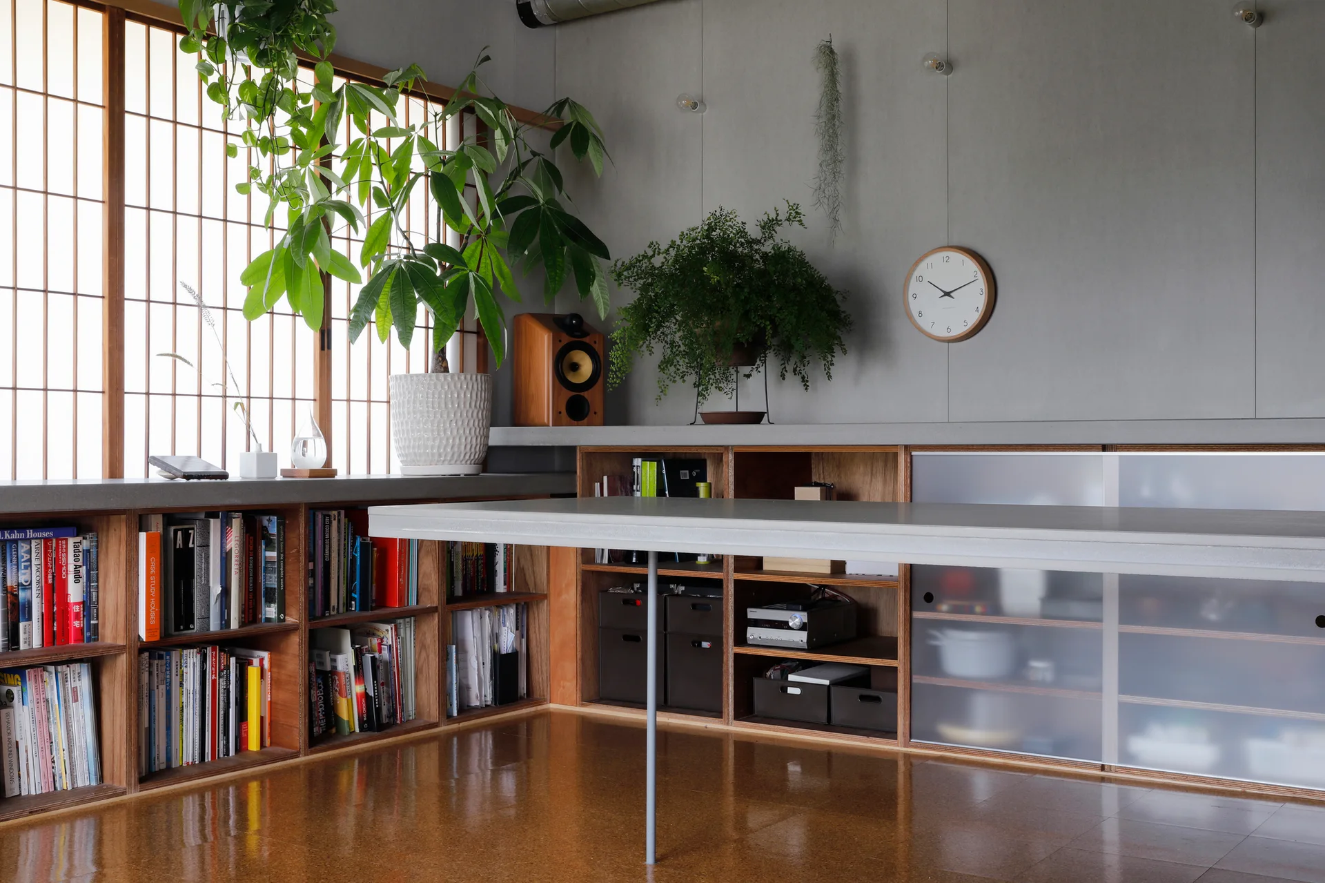 A modern workspace features low wood built-in bookshelves, a long grey table, a grid-patterned window, and potted plants against a grey wall.