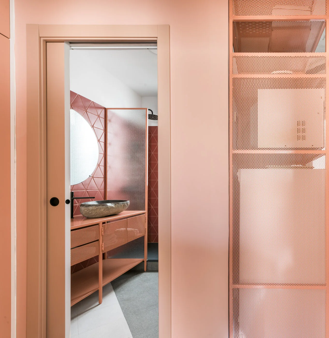 A monochromatic pink bathroom viewed through an open doorway, featuring a pink perforated metal vanity with a stone vessel sink and a round mirror.