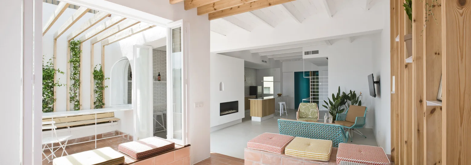 An open-concept living area with white walls and exposed ceiling beams transitions into a sunlit terracotta patio with climbing vines.