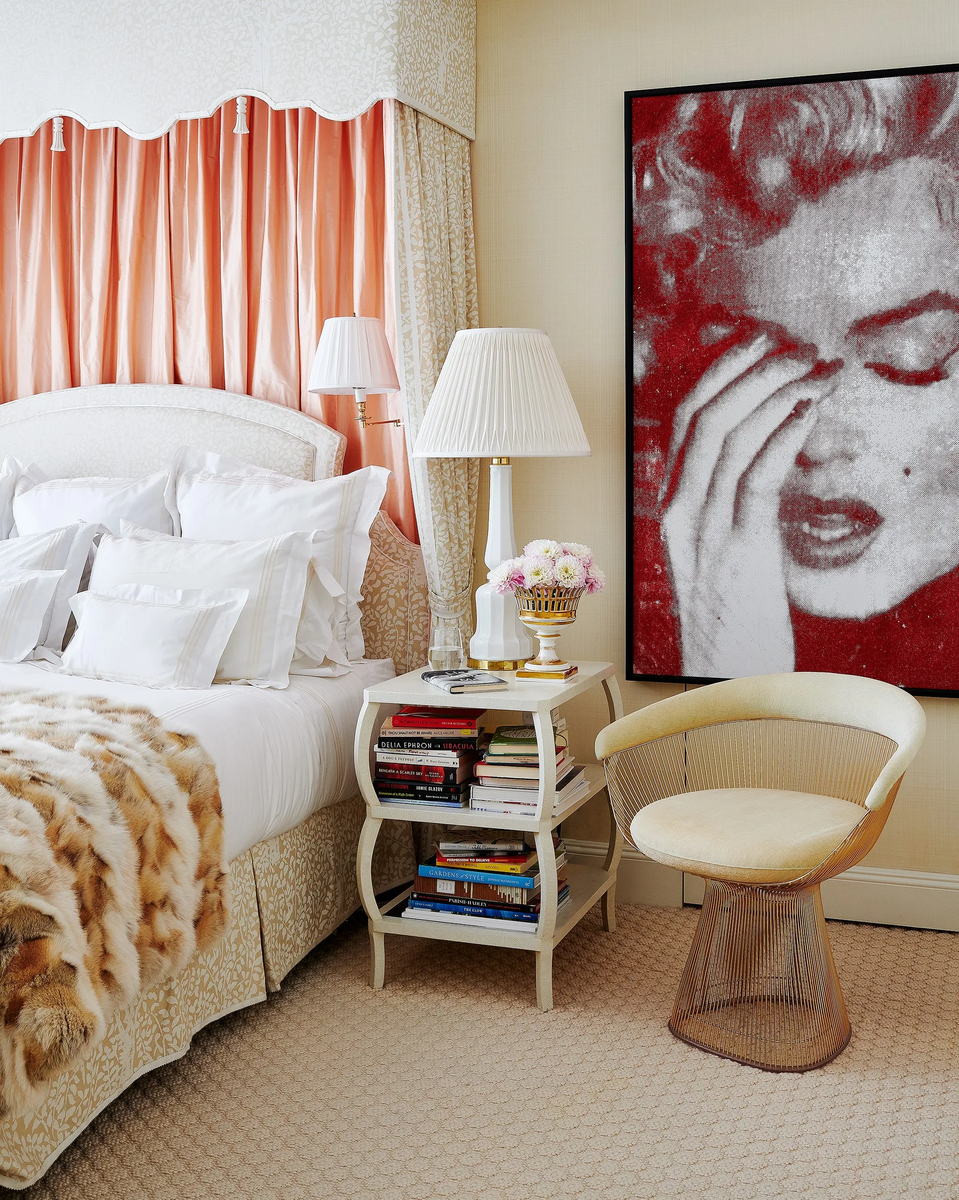 A bedroom corner features a bed with white linens, a peach curtain backdrop, a two-tier book-filled nightstand, a gold wire chair, and red wall art.