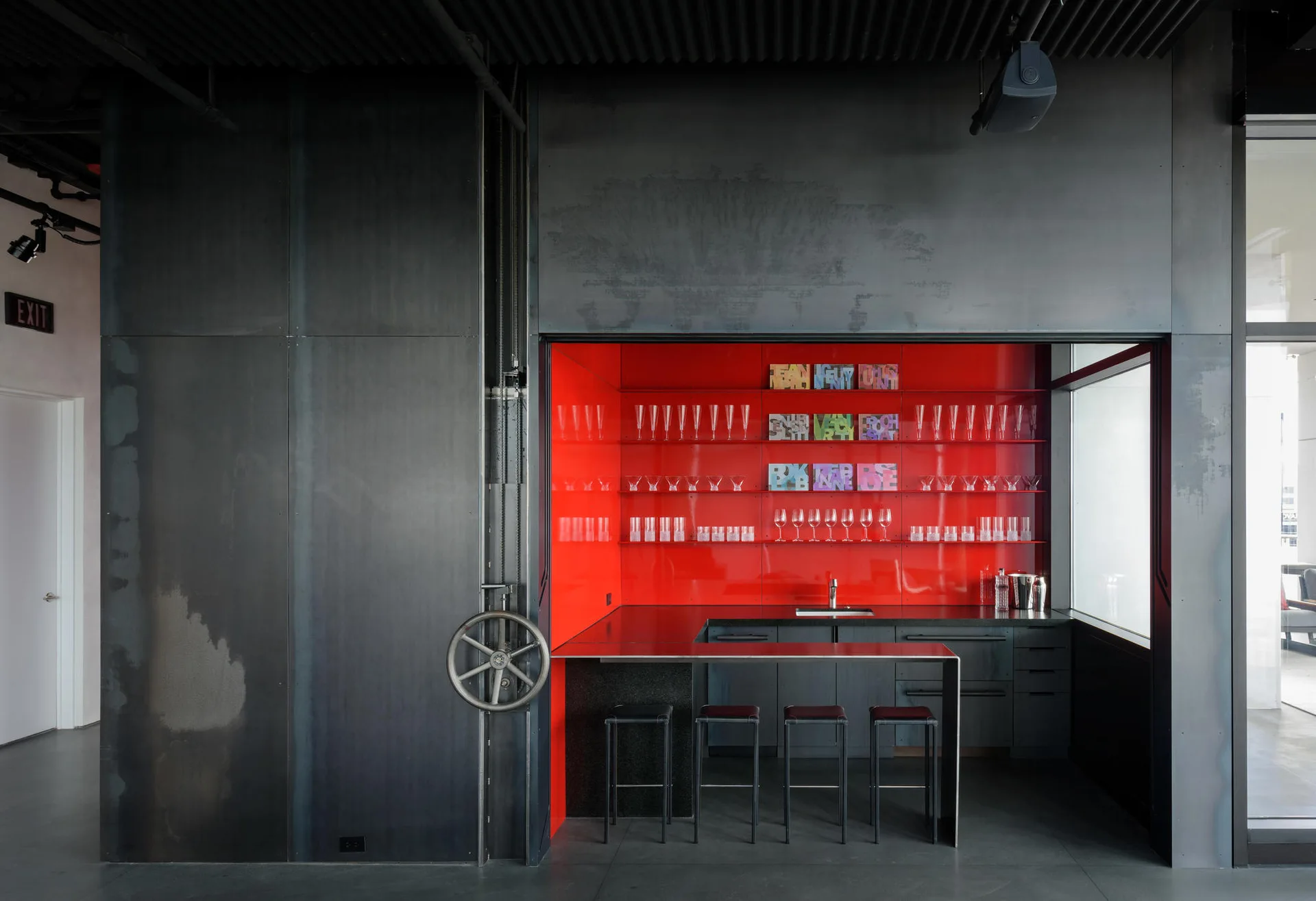An industrial bar area featuring dark steel wall panels, an integrated hand wheel, and a recessed bar niche with bright red walls and glass shelving.