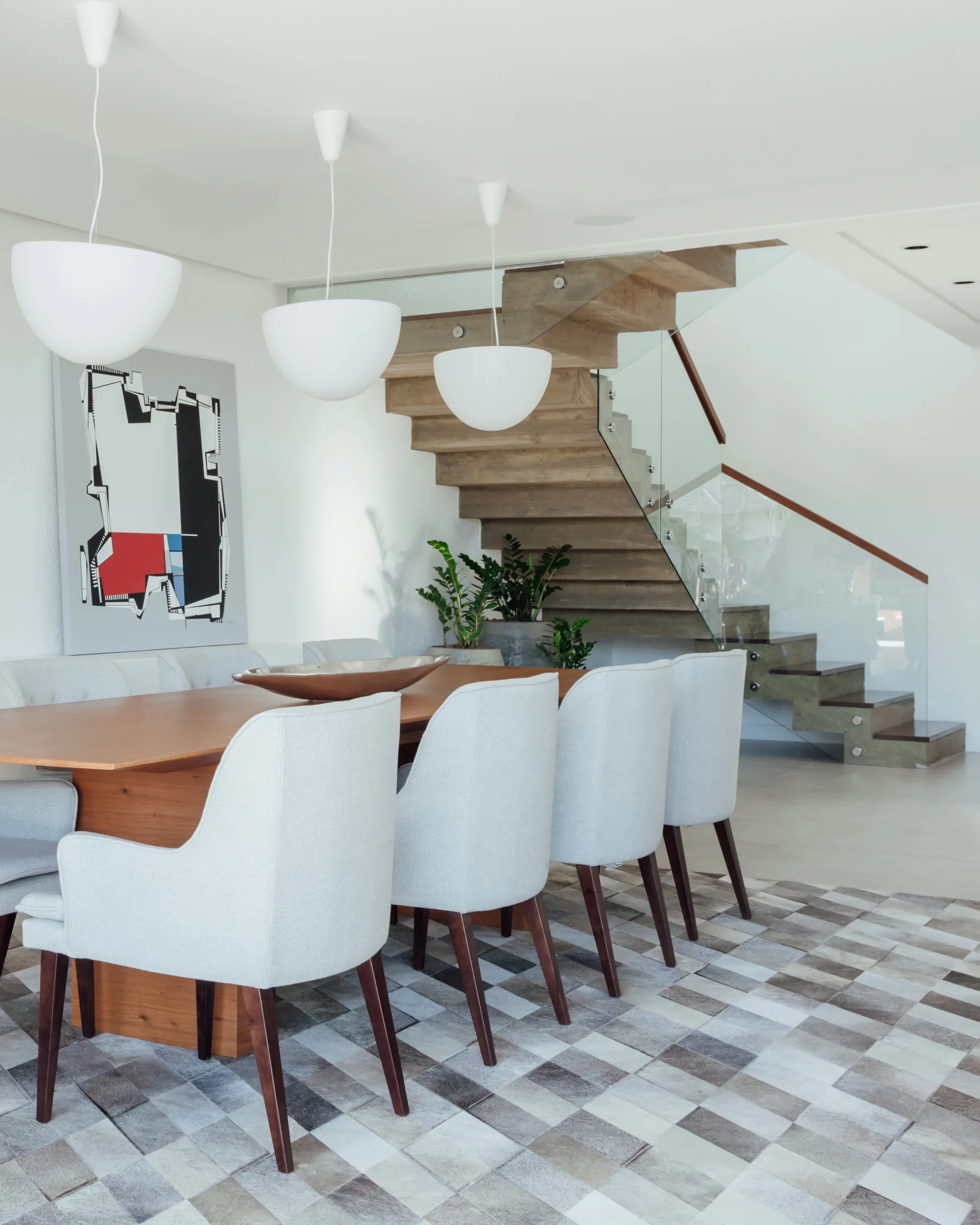 A modern dining area with a wood table, light gray upholstered chairs, and three white bowl pendant lights near a floating staircase.