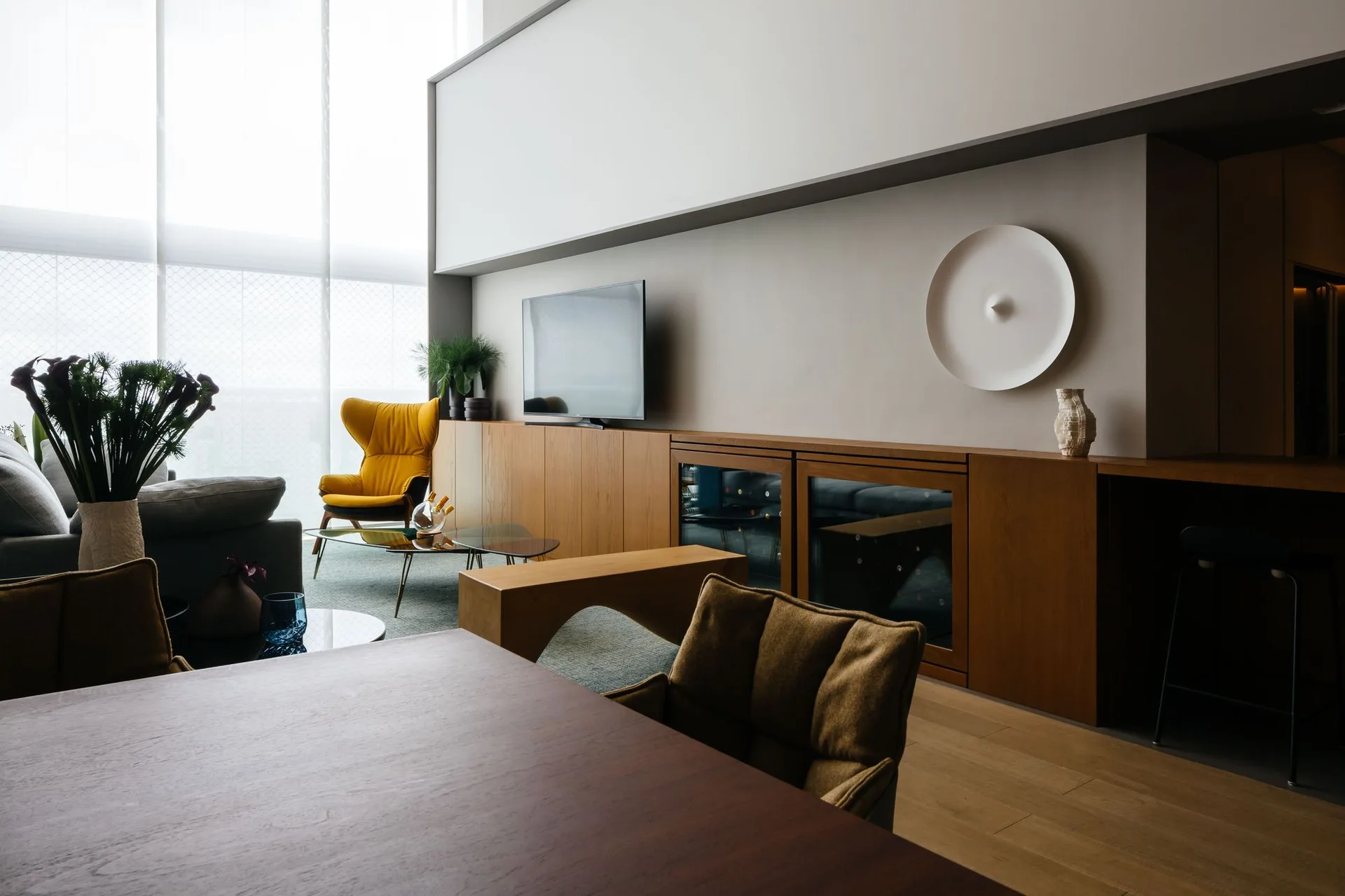 A modern living room features a long wooden media console, a yellow wingback armchair, a glass coffee table, and a large window with sheer white curtains.