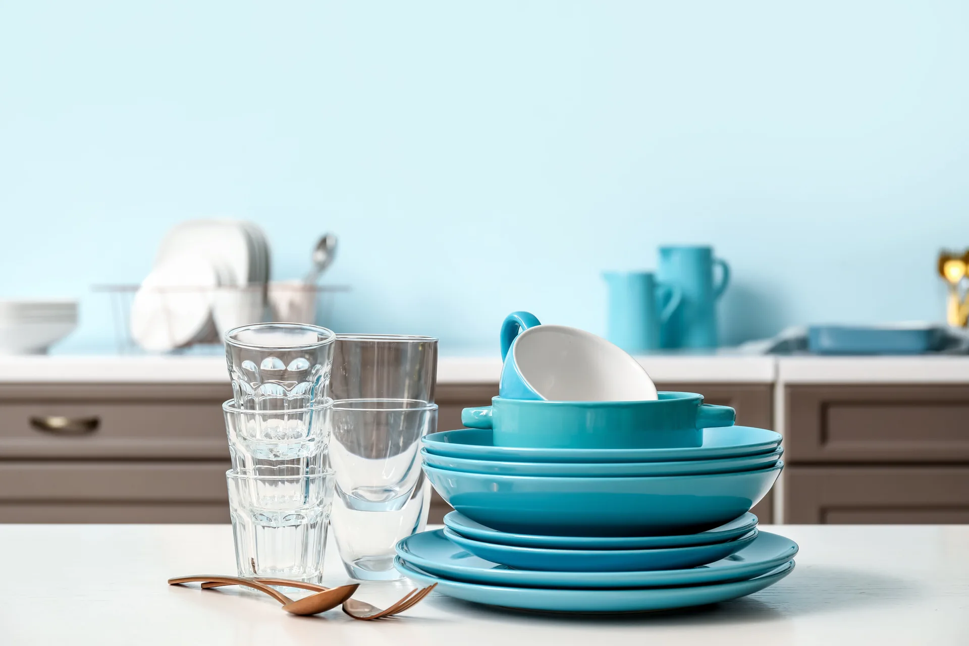 A stack of bright blue ceramic plates and bowls sits next to clear drinking glasses and bronze cutlery on a white tabletop.